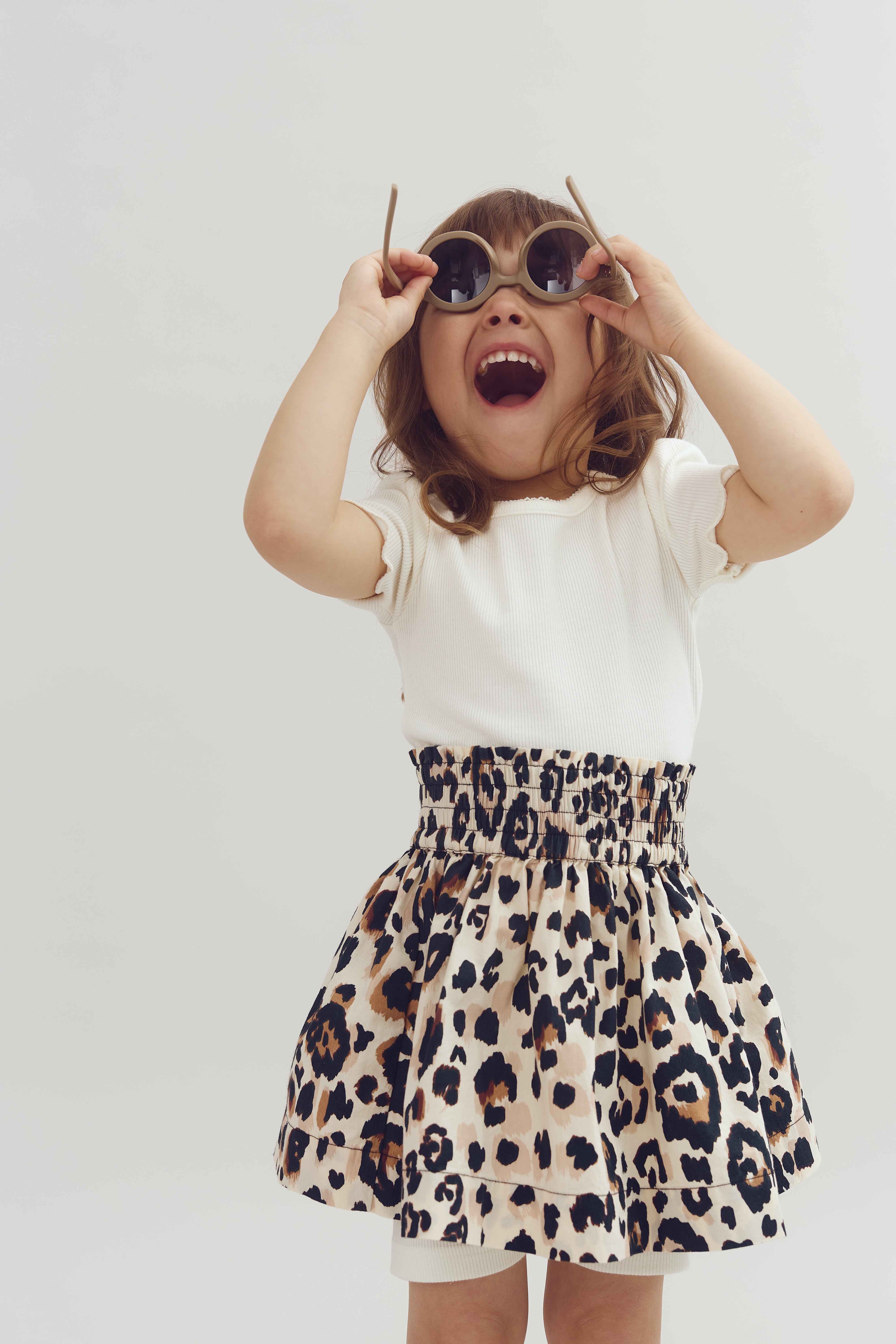 Child wearing round sunglasses and a wide smile, lifting the shades slightly. Dressed in a white shirt and leopard-print skirt, set against a plain, light grey background.