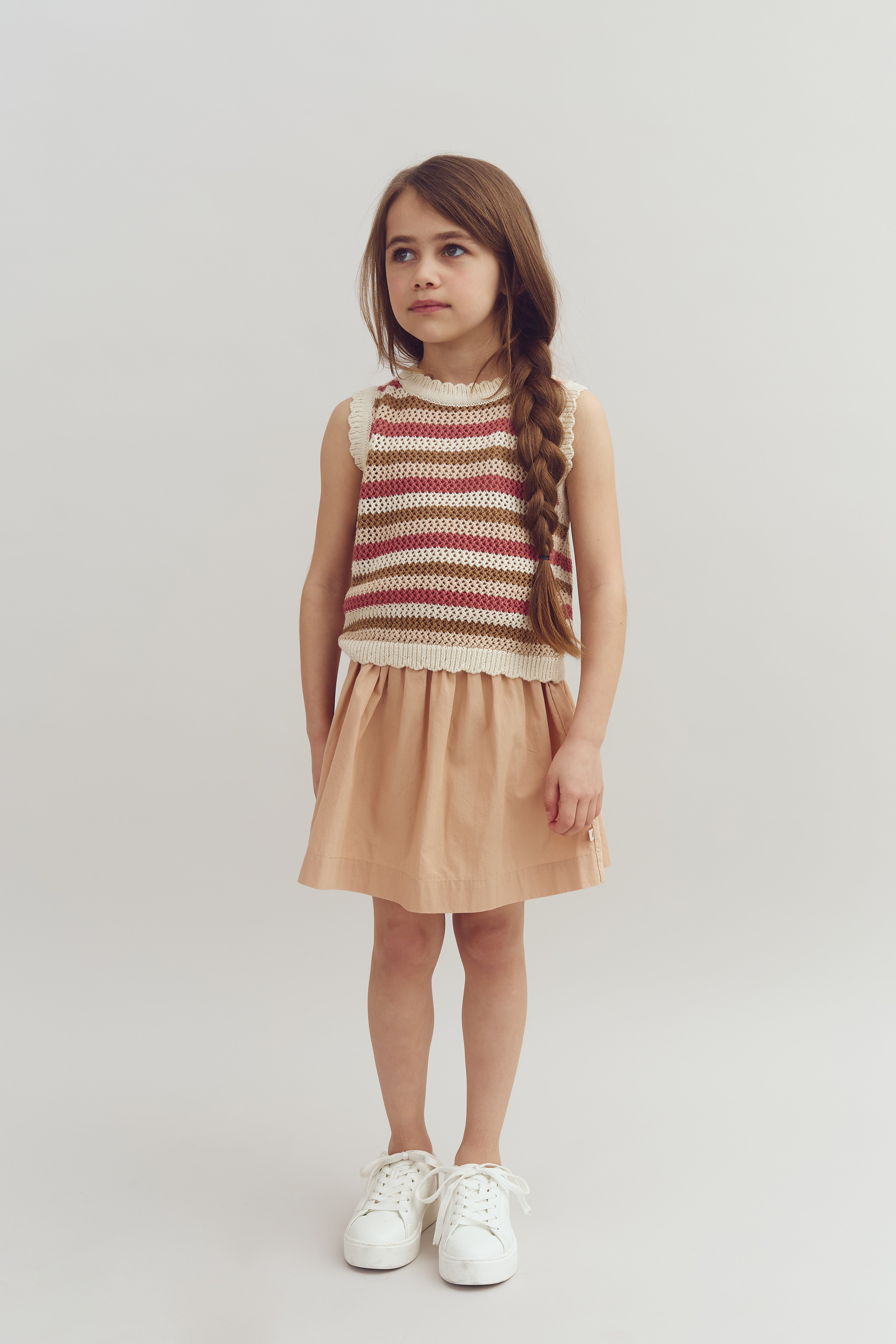 A young girl stands wearing a striped sleeveless top and a beige skirt, with a long braid over her shoulder, against a plain, light-colored background.