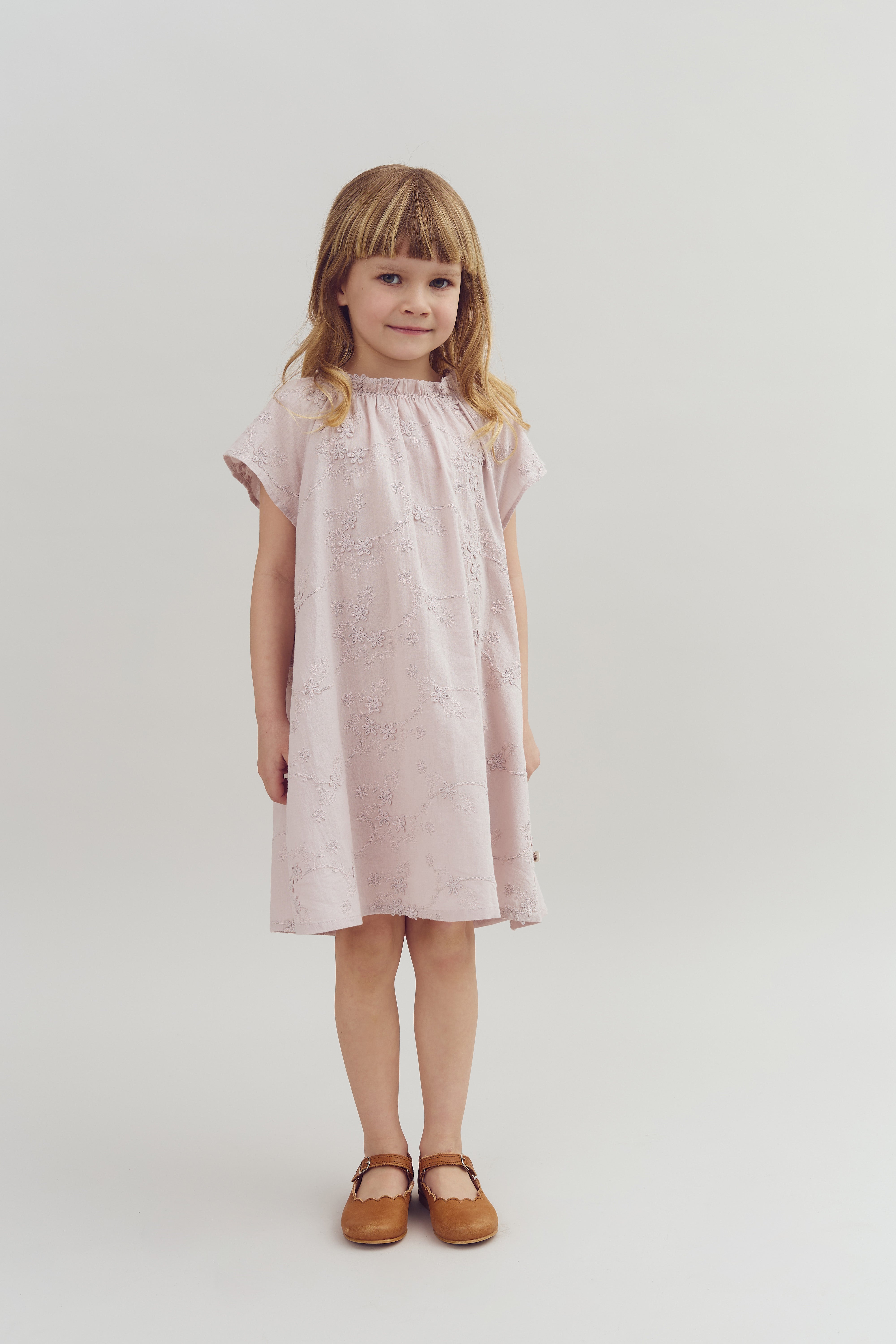 A child is standing, wearing a light pink dress with floral patterns and brown shoes, in a plain white studio setting.
