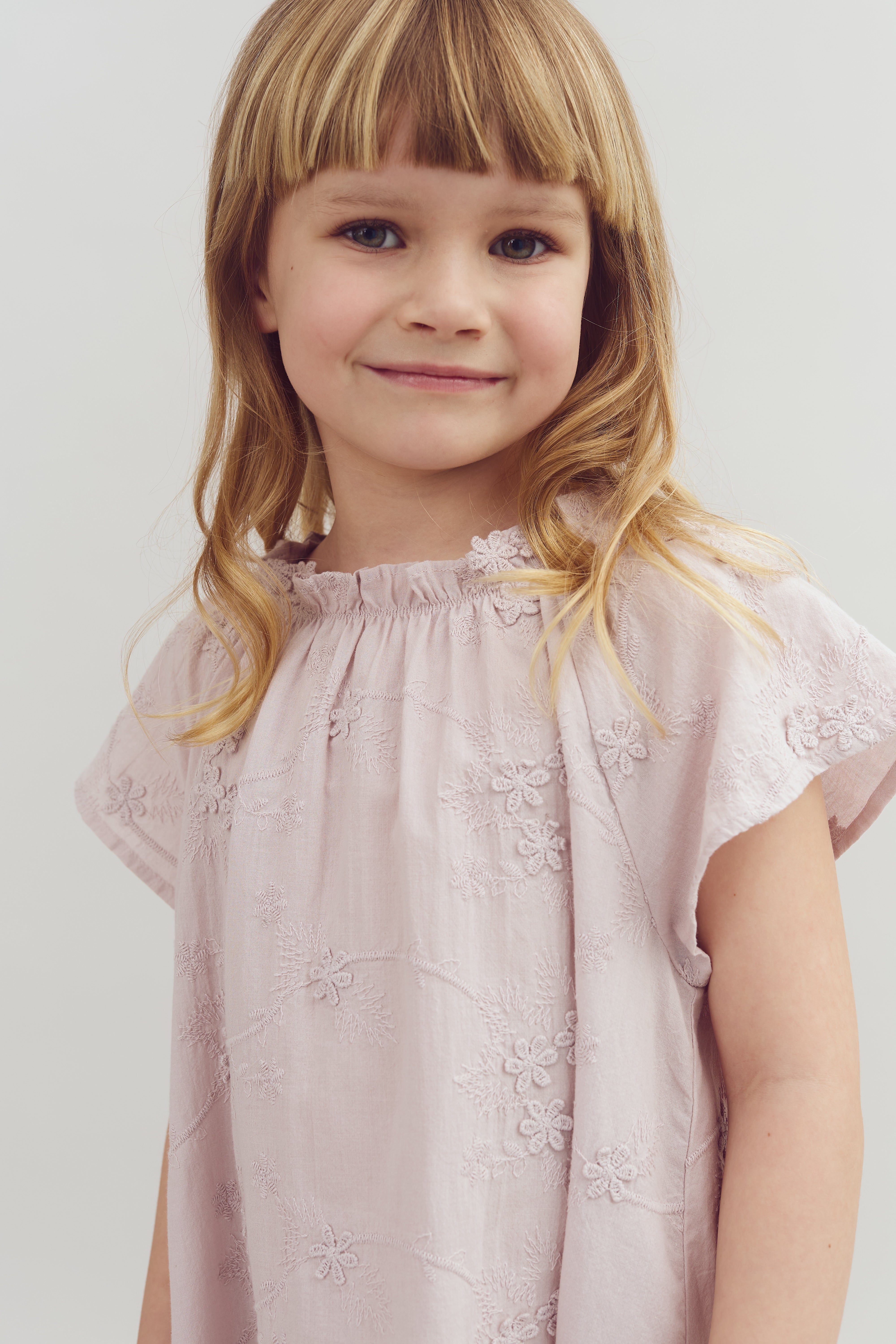 A young child smiles gently, wearing a light pink embroidered dress with floral patterns, in front of a plain, light-colored background.