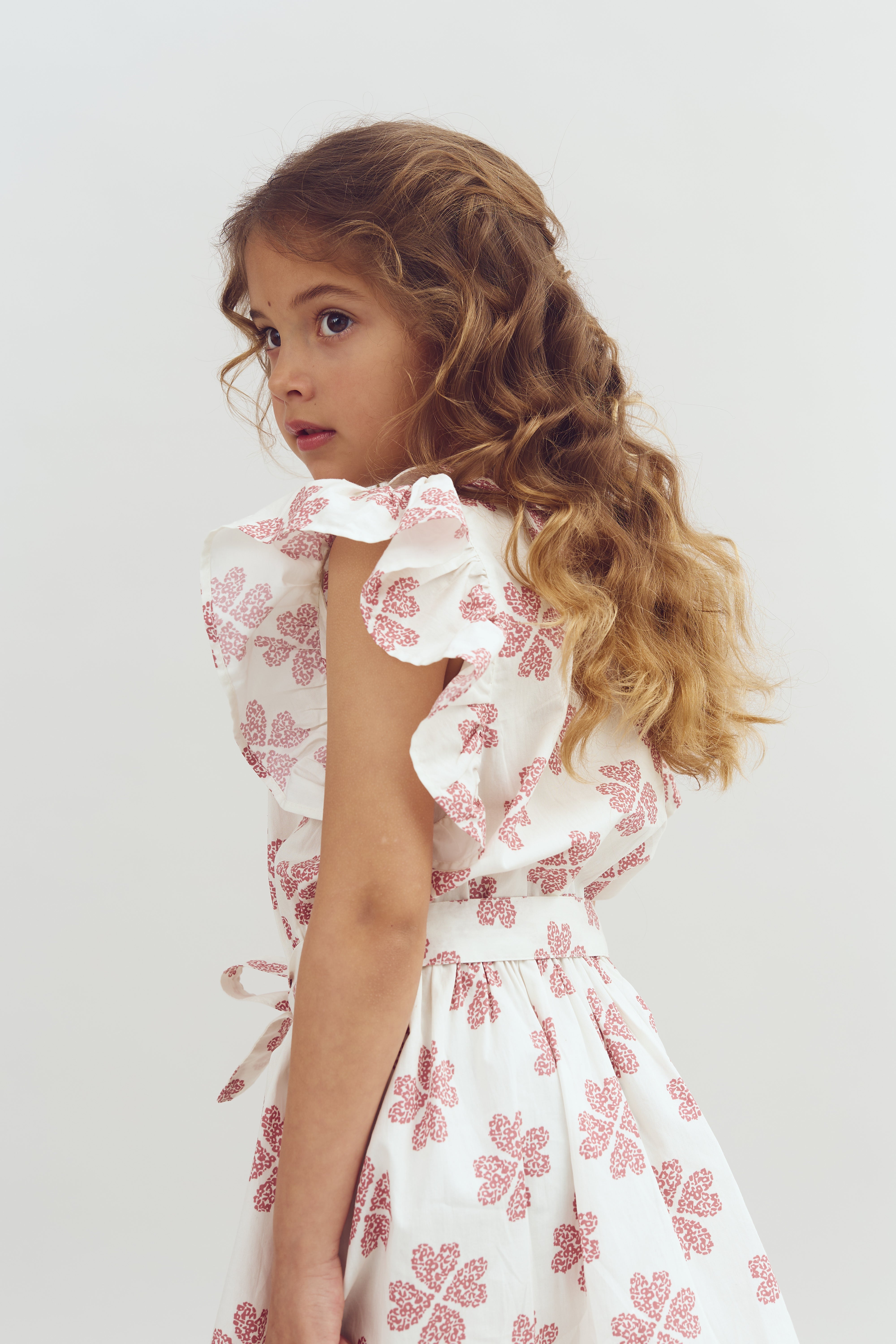 A young girl with long, wavy brown hair stands sideways, wearing a white dress with red floral patterns, against a plain, light background.
