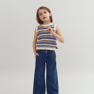 A child stands with one hand in a pocket and the other raised, wearing a striped sleeveless top and wide-leg jeans, against a plain, light background.