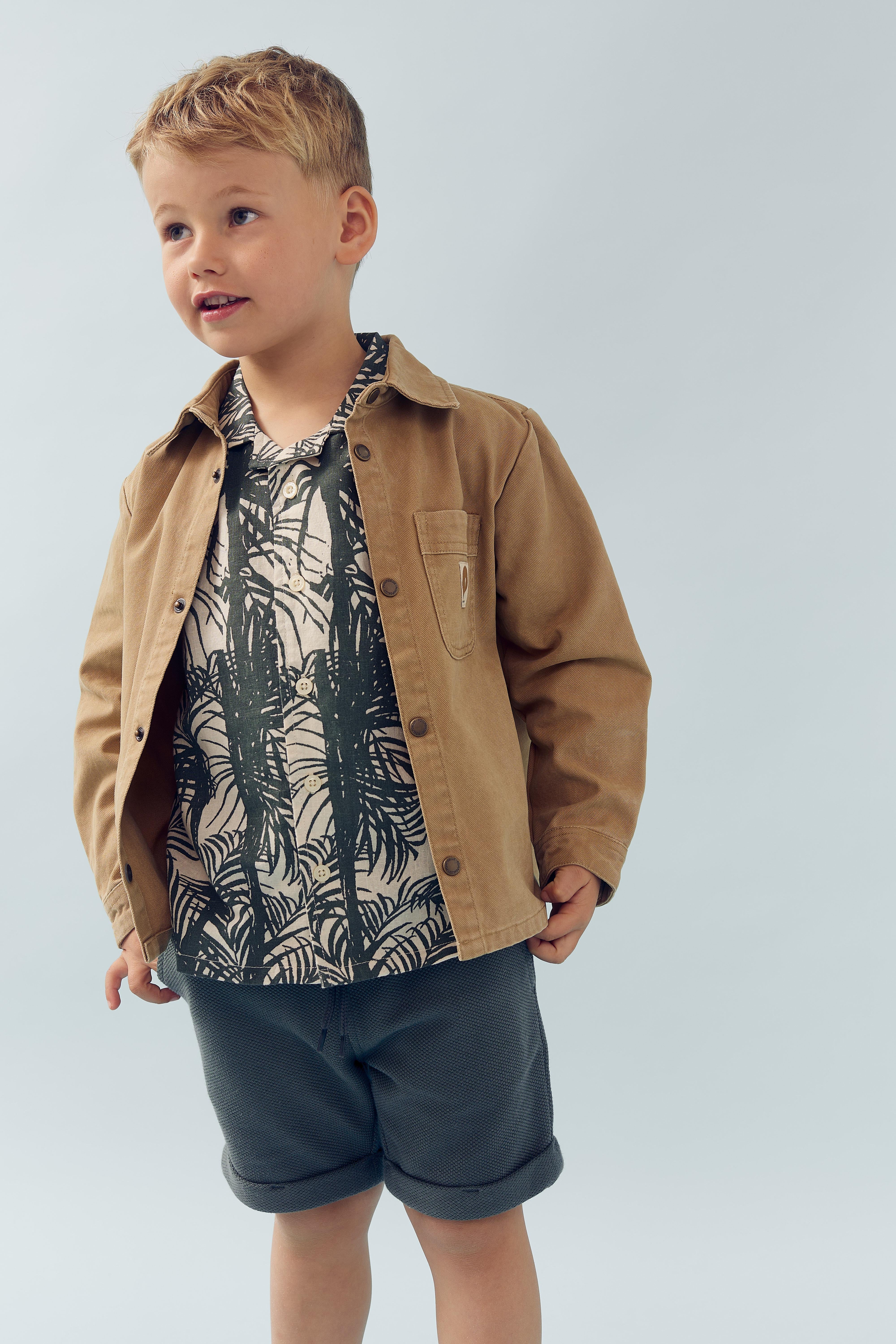 Een jongen draagt een beige jas en een overhemd met palmbladprint, gecombineerd met korte broek. Hij staat tegen een neutrale, lichtblauwe achtergrond en kijkt omhoog.