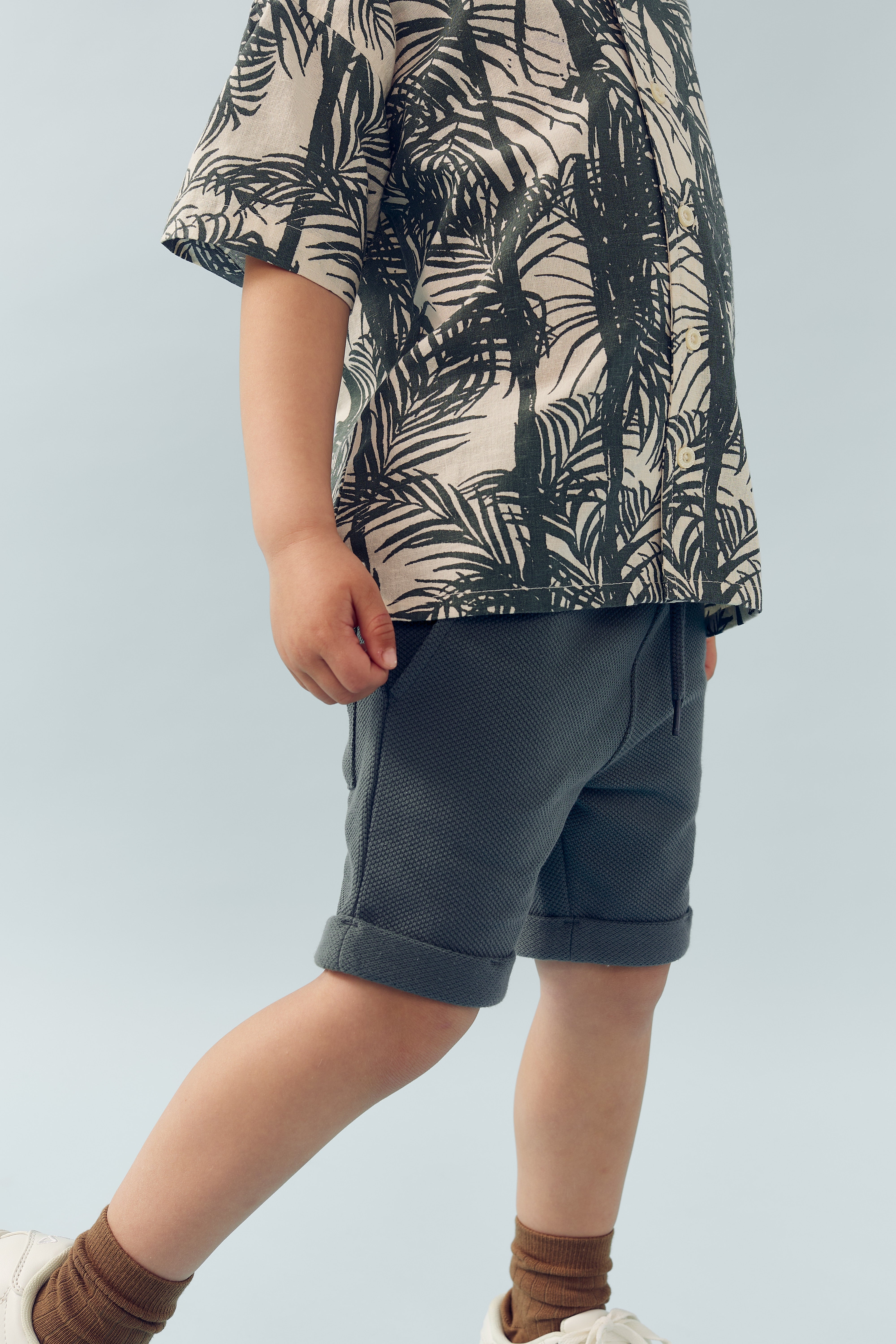 Kind draagt een shirt met palmbomenprint, grijze korte broek en bruine sokken in witte schoenen, staande tegen een lichtblauwe achtergrond.
