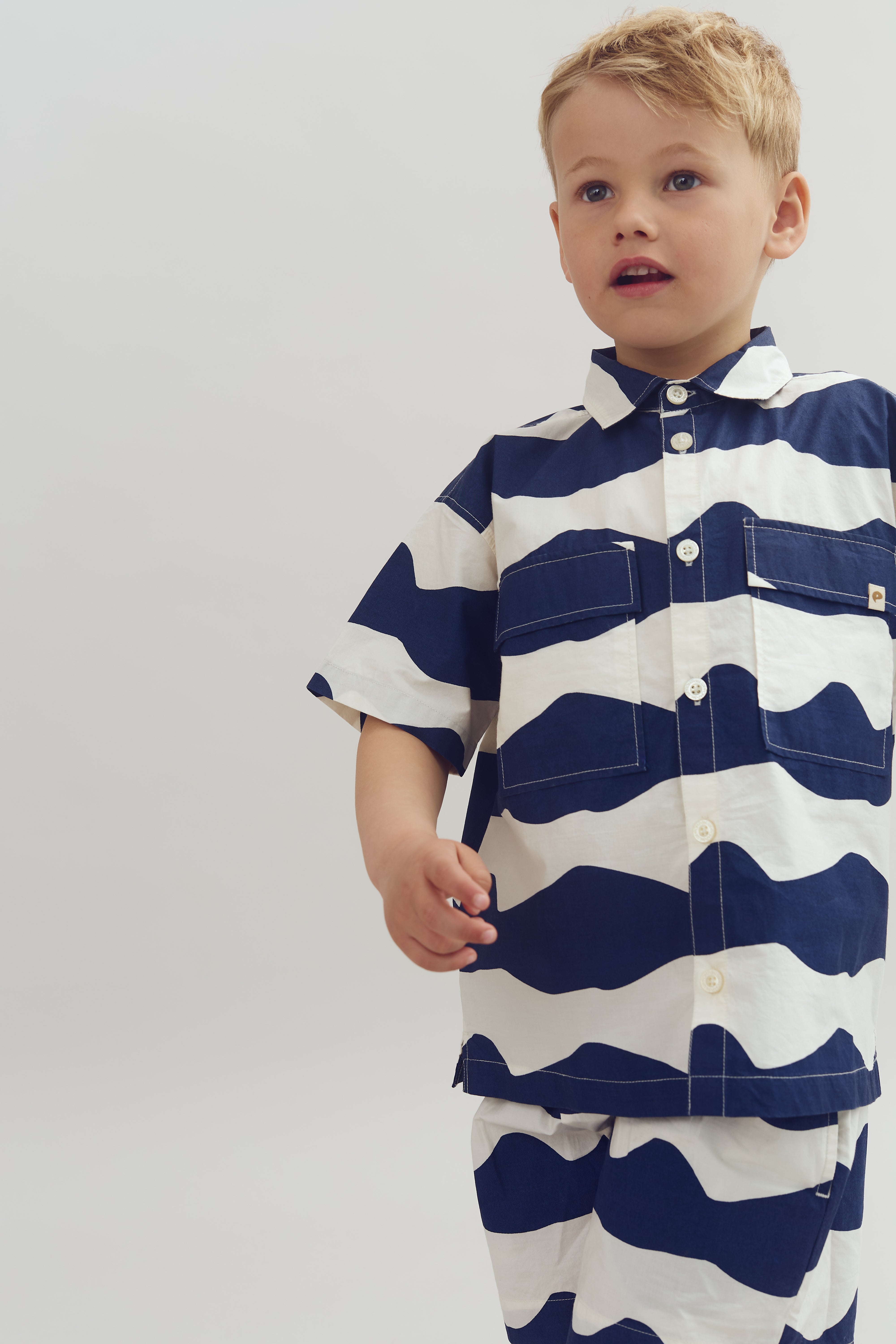 Een jongen draagt een blauw-wit gestreept pak terwijl hij naar rechts kijkt, tegen een eenvoudige, lichte achtergrond.