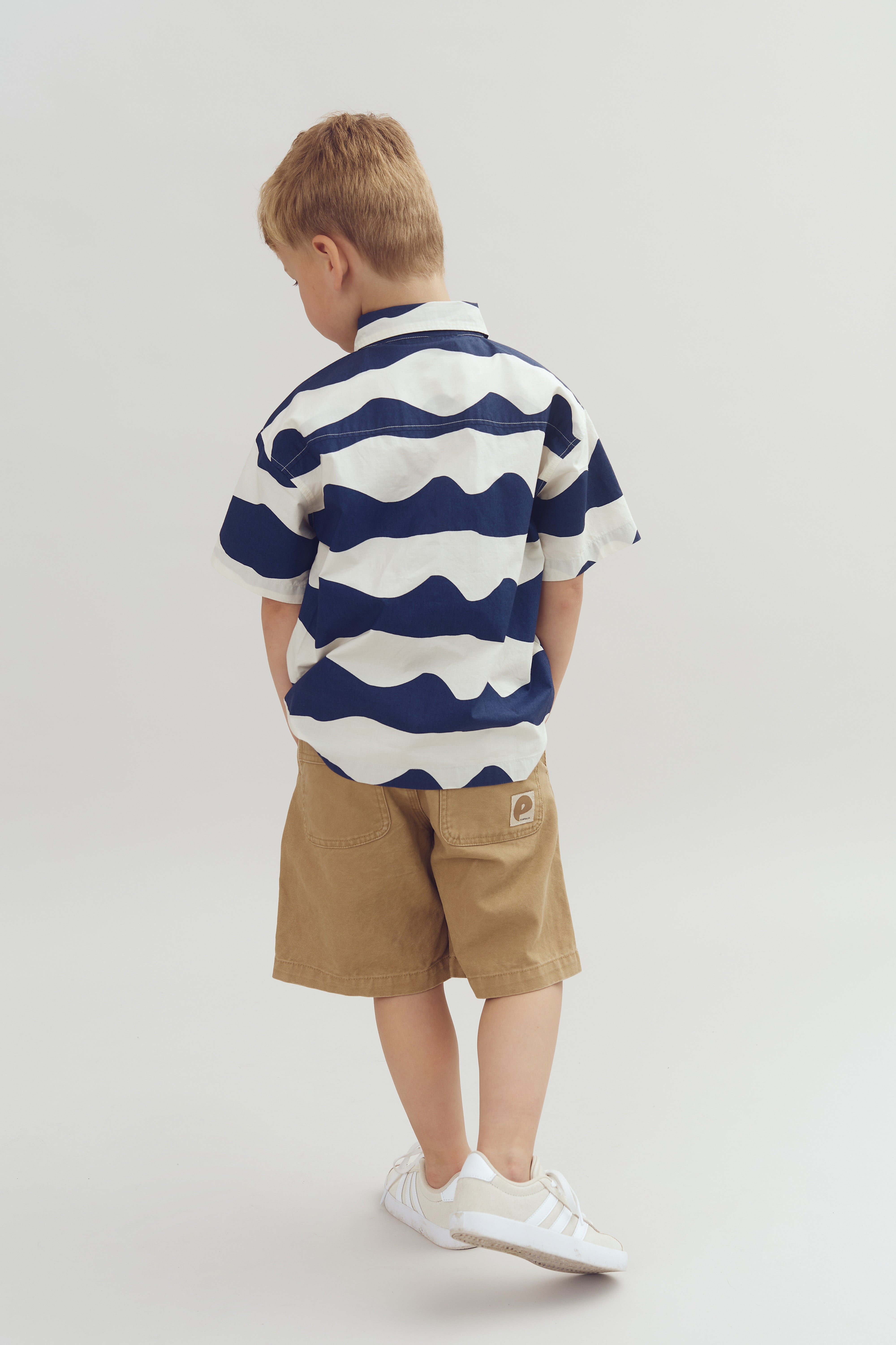 Een kind staat met rug naar de camera, draagt een gestreept T-shirt en bruine shorts. De omgeving is een neutrale, lichtgekleurde achtergrond. Er is geen tekst zichtbaar.