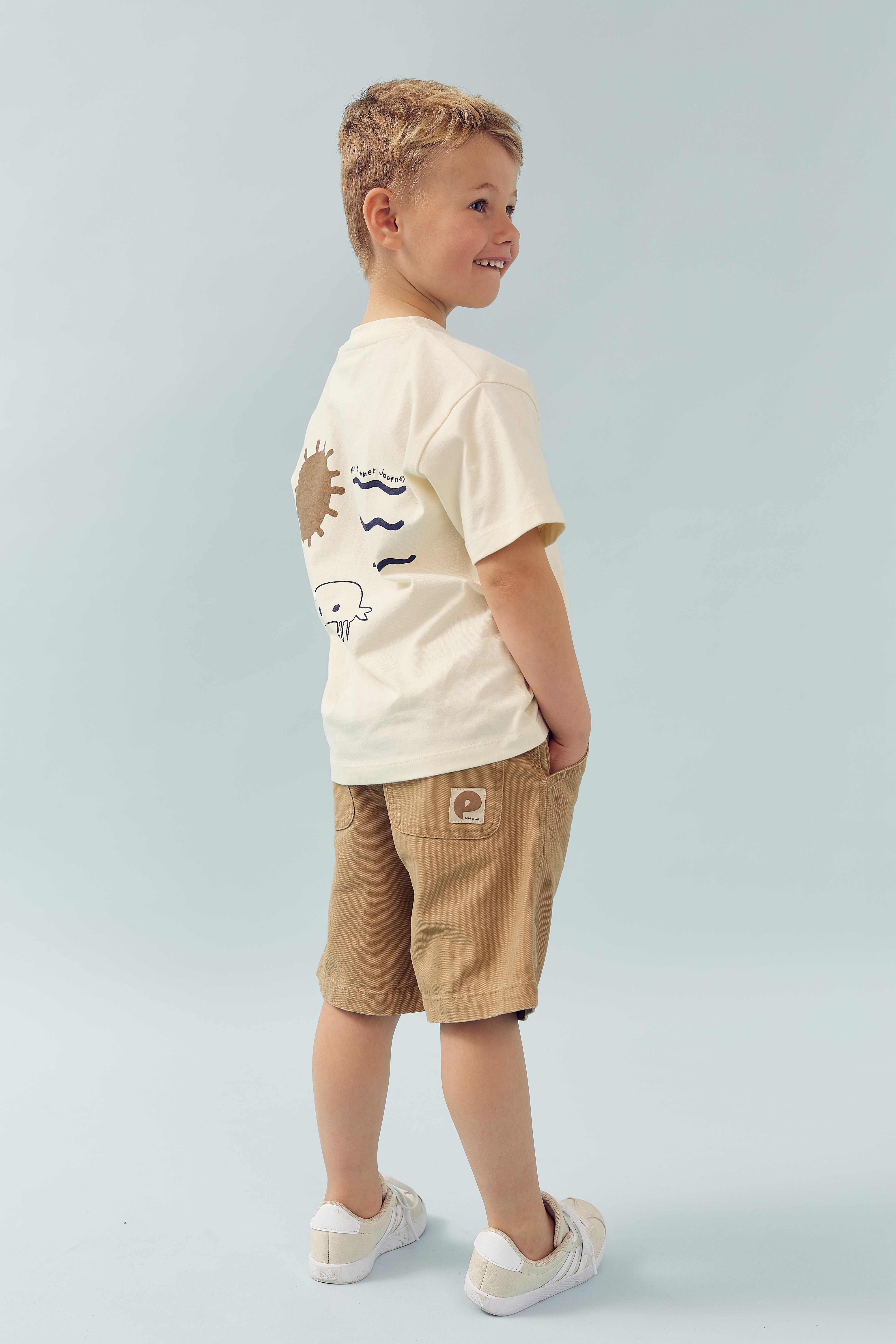 Een jongen draagt een beige T-shirt met tekeningen en bruine korte broek. Hij staat zijwaarts met zijn handen in de zakken, op een lichtblauwe achtergrond. Op het T-shirt staat "no sugar, thanks."