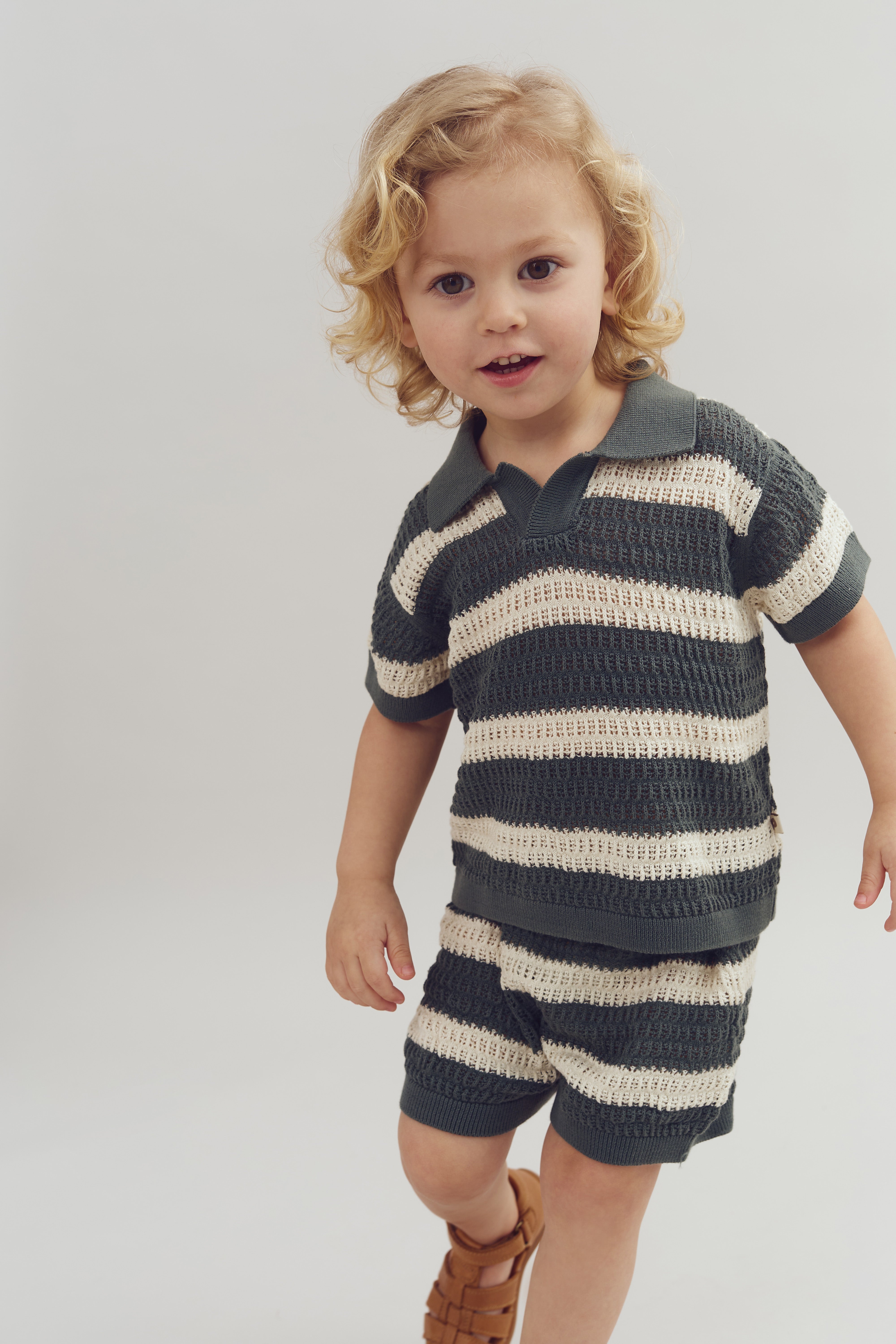 A child with curly hair stands, wearing a striped knit outfit and sandals, against a plain background. They are slightly smiling, with one hand up, conveying a playful demeanor.
