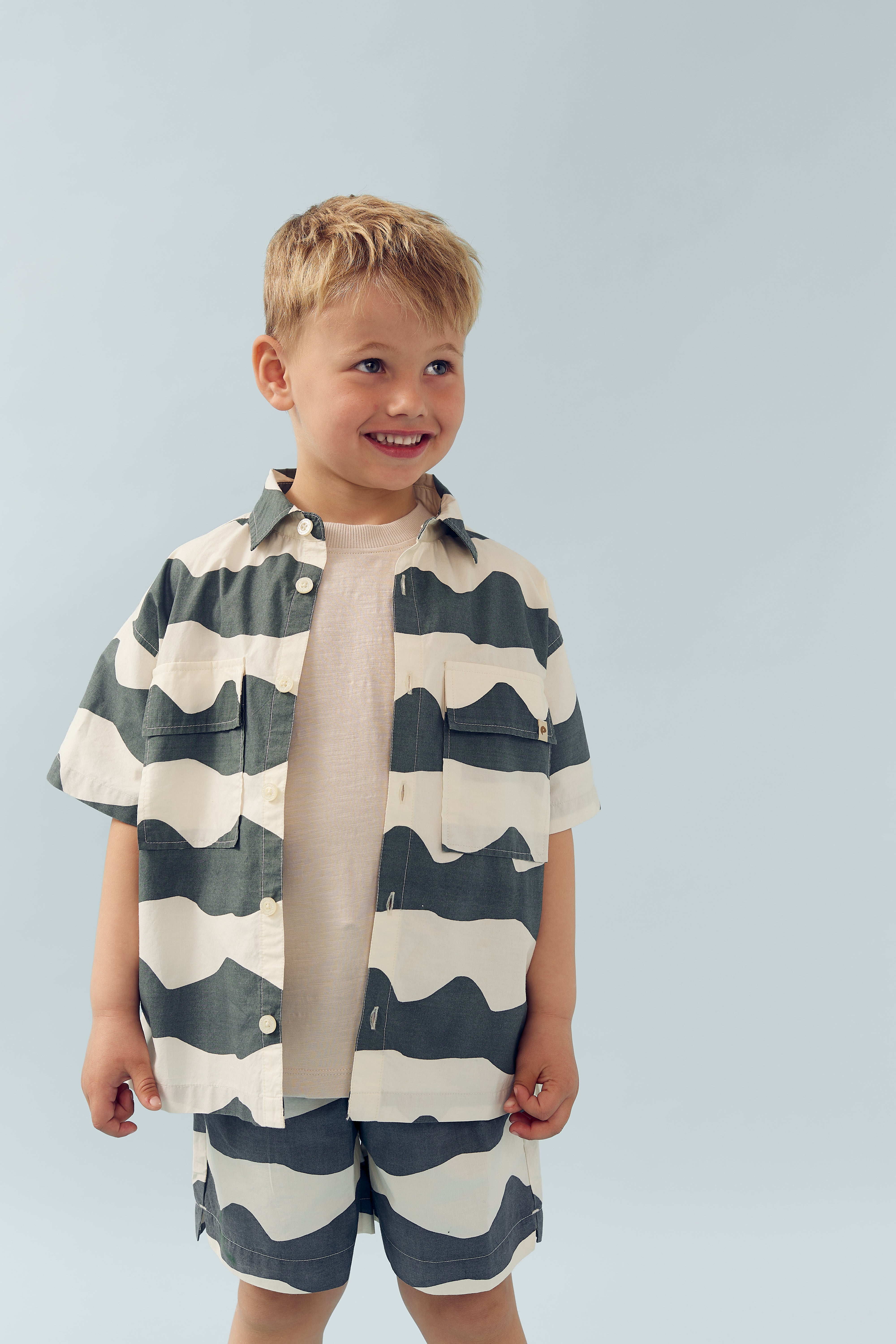 Een jongen draagt een gestreept overhemd en korte broek, glimlachend binnen een minimalistische, lichte achtergrond.