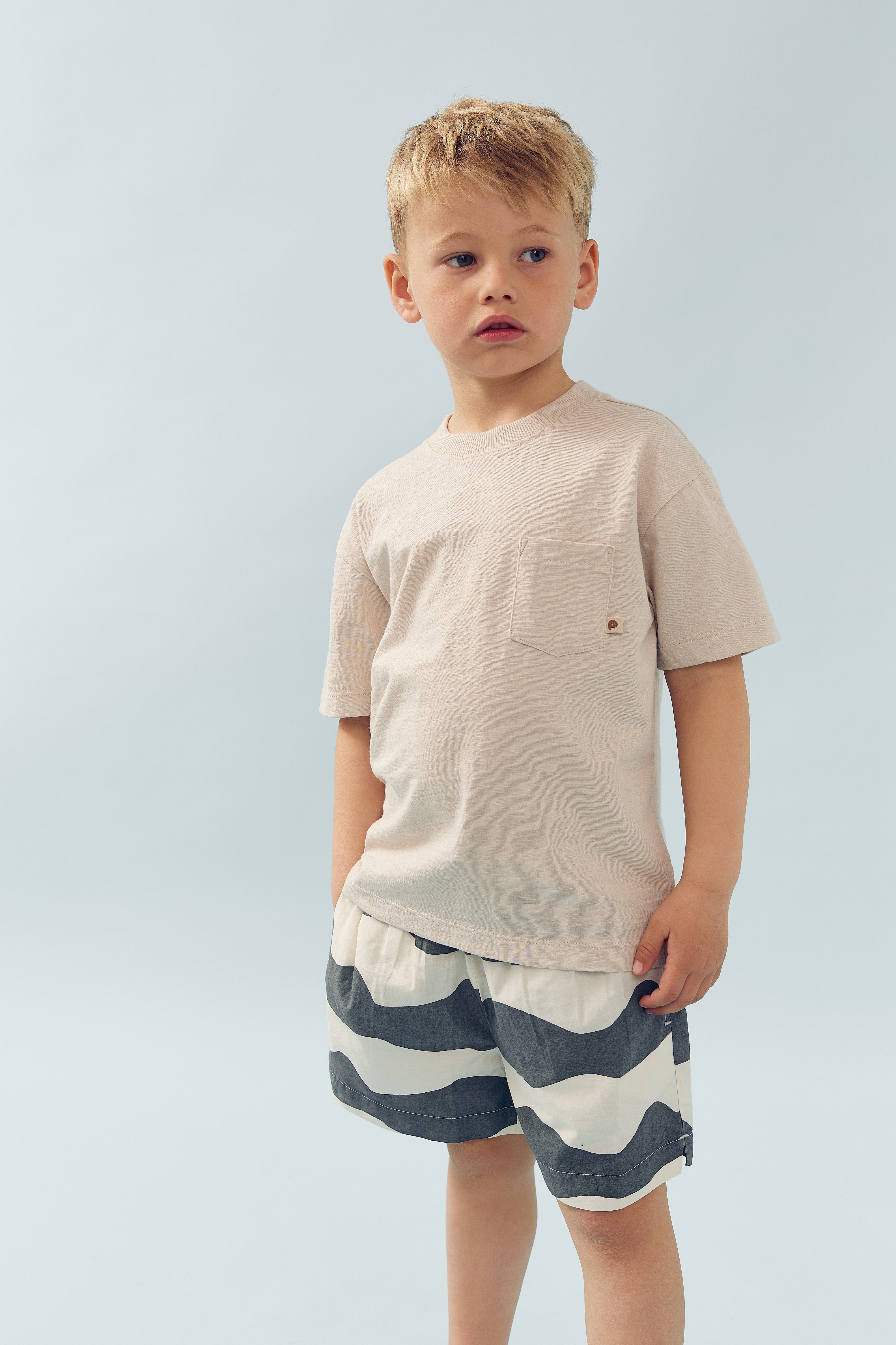 A young boy stands wearing a light beige T-shirt and striped shorts, looking off to the side. The background is a plain light blue.