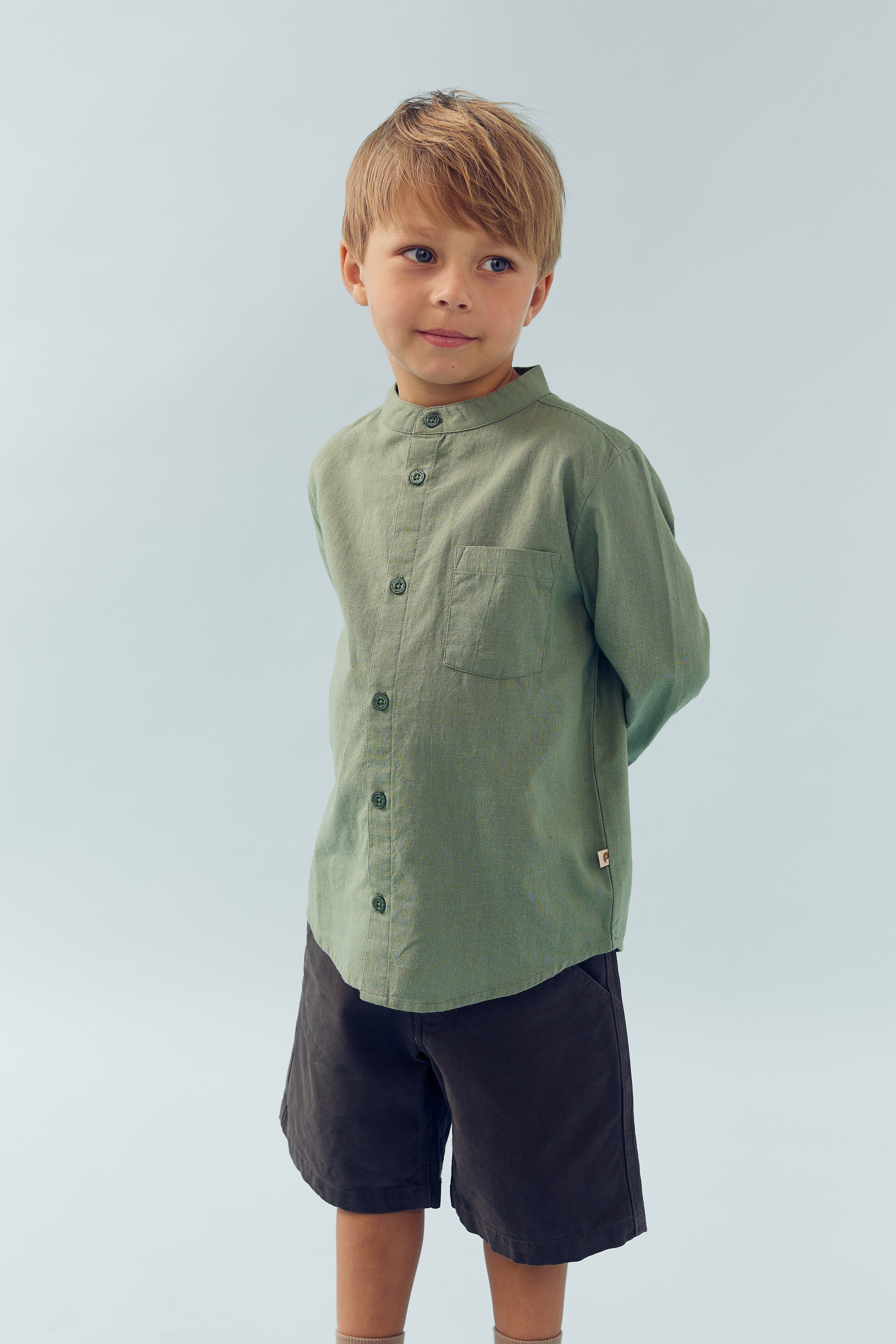 Een jongen staat met zijn handen achter zijn rug, gekleed in een groene blouse en donkerbruine shorts, tegen een effen, lichtblauwe achtergrond.