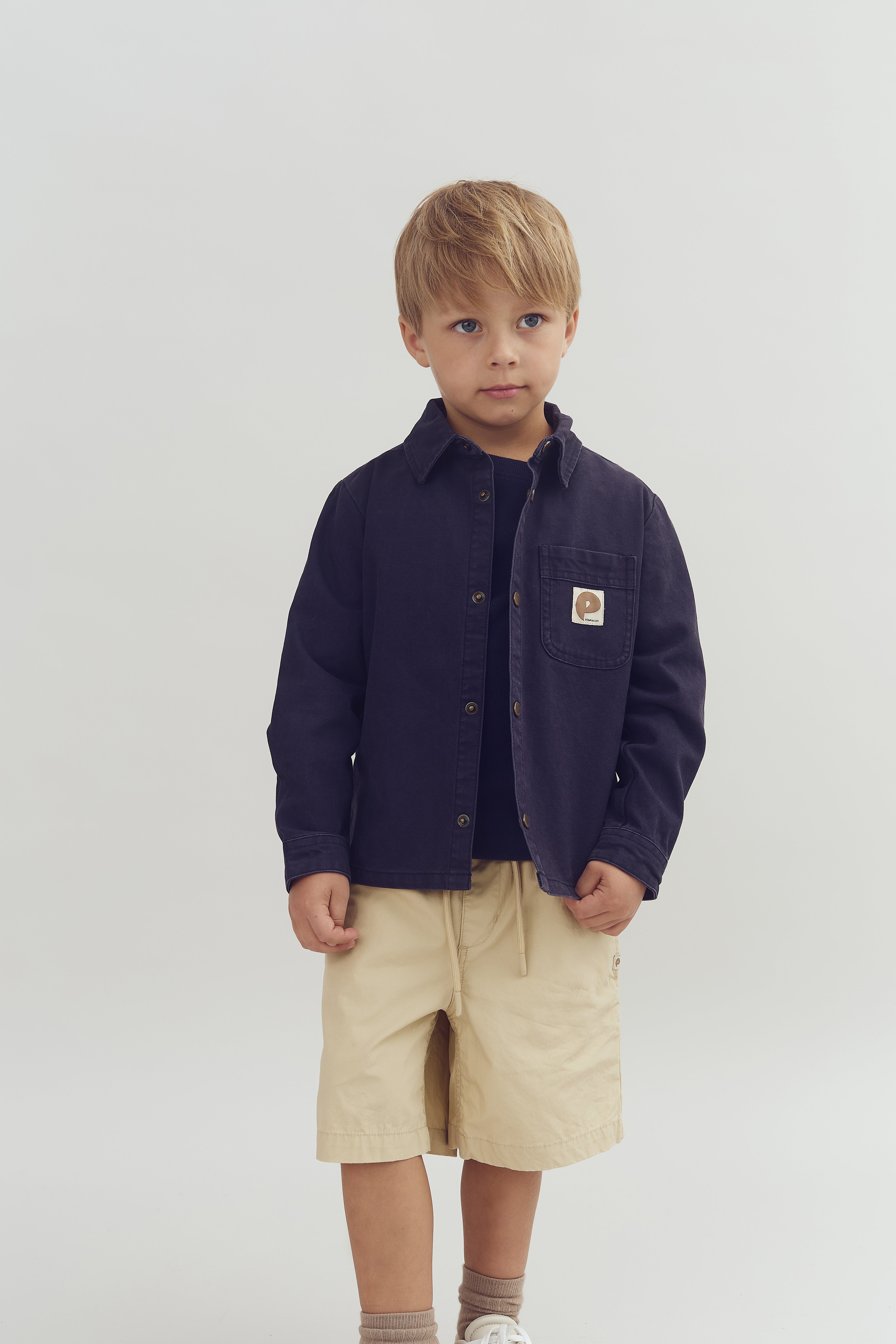 Een jonge jongen staat stil, gekleed in een donker overhemd met een embleem op de borst en beige korte broek, tegen een lichte, egale achtergrond.