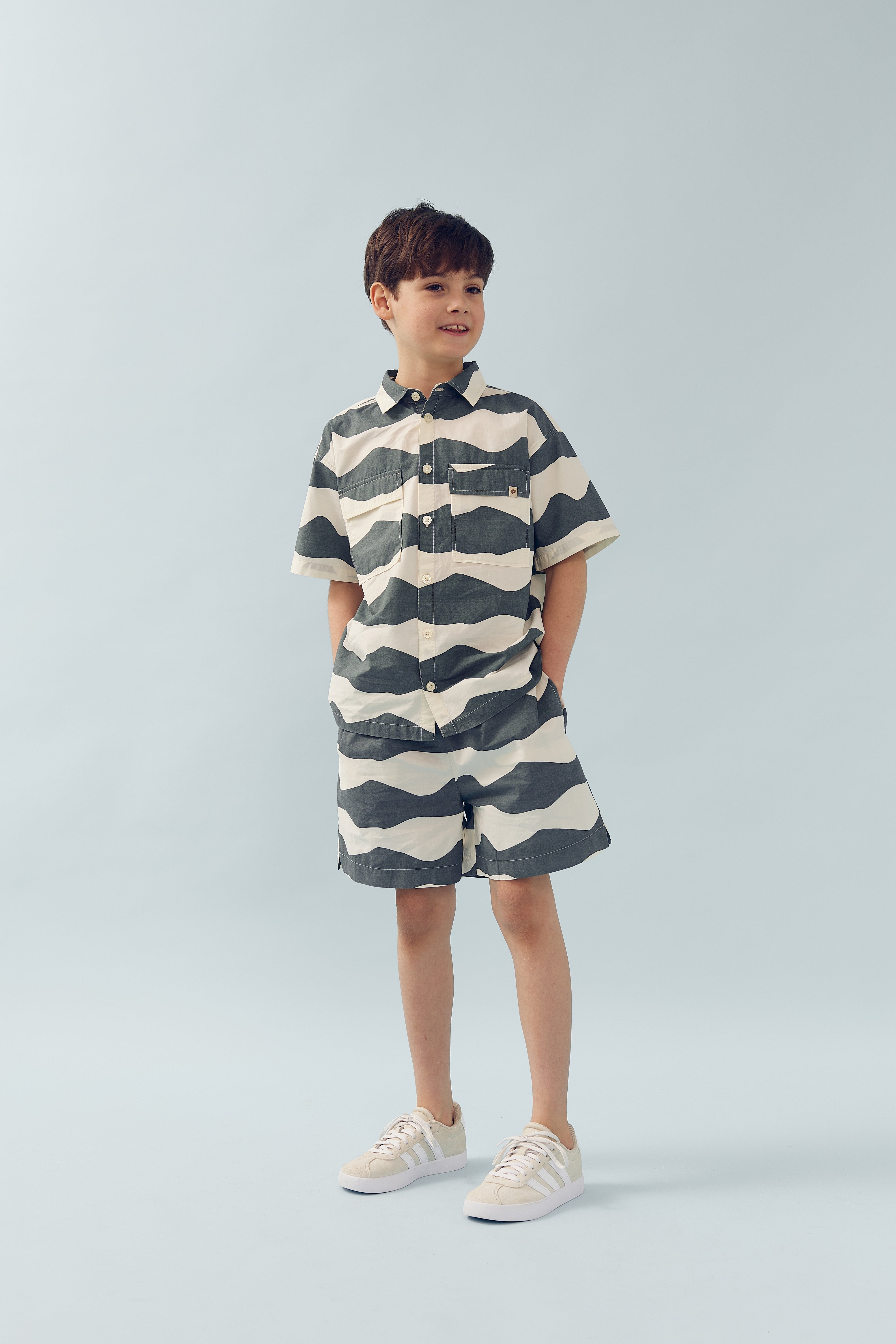 A child stands casually with hands in pockets, wearing a matching wavy-striped shirt and shorts set. The background is a plain light blue, creating a clean, minimalistic environment.