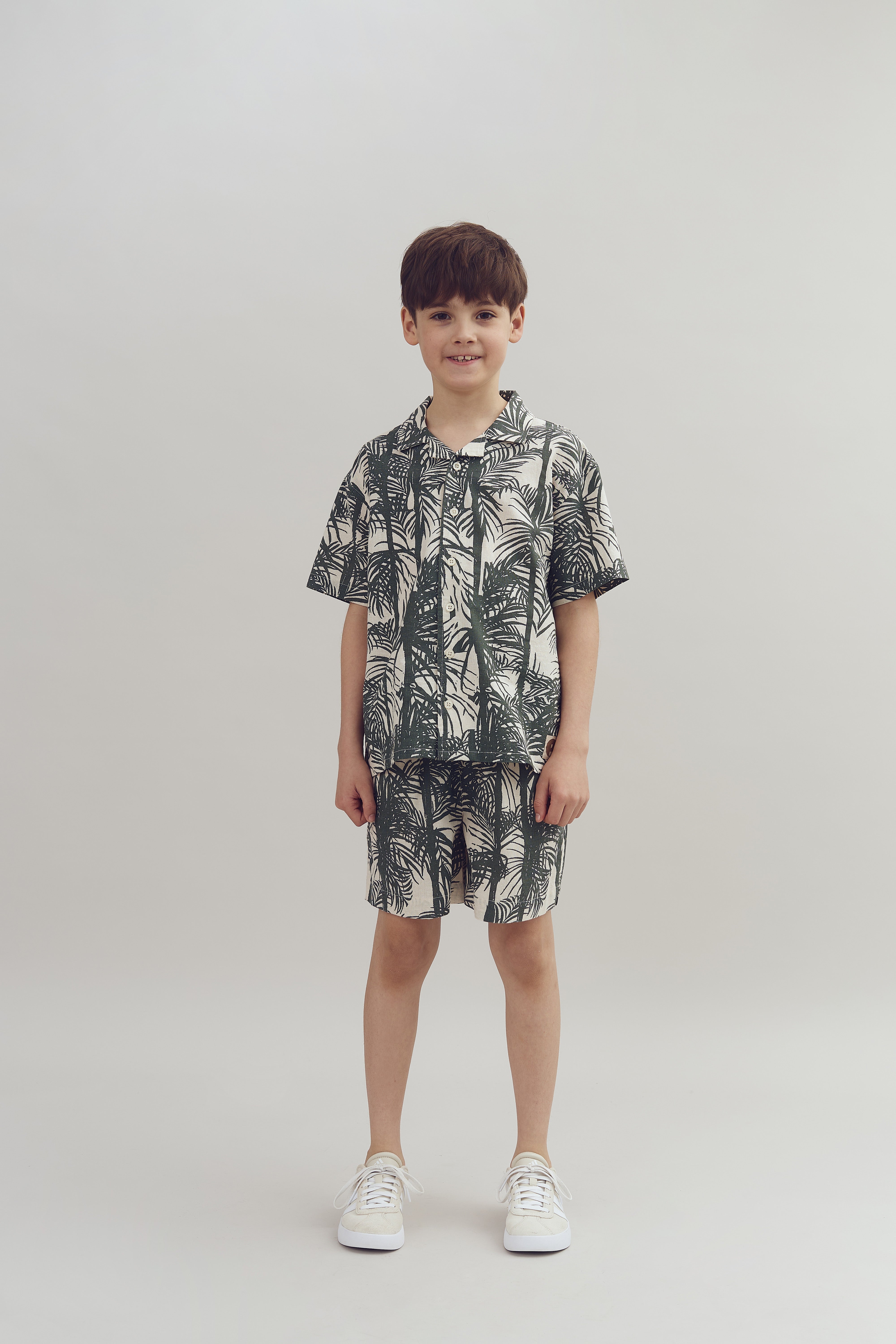 A child stands smiling, wearing a matching set of tropical-patterned shirt and shorts, paired with white sneakers, against a plain gray background.