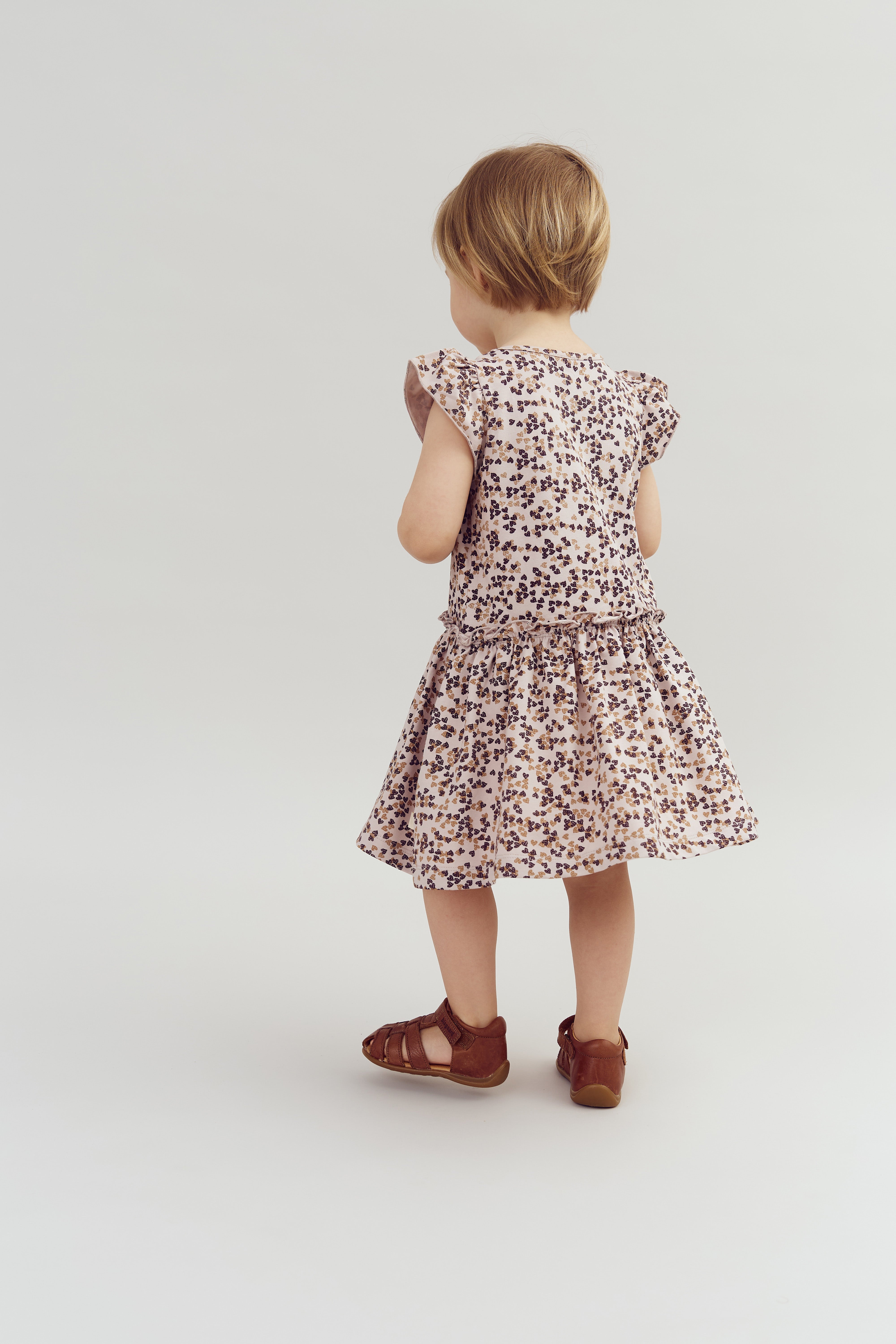 Een klein kind in een bloemenjurk staat met de rug naar de camera, in een neutrale, witte ruimte. Het kind draagt bruine sandalen.