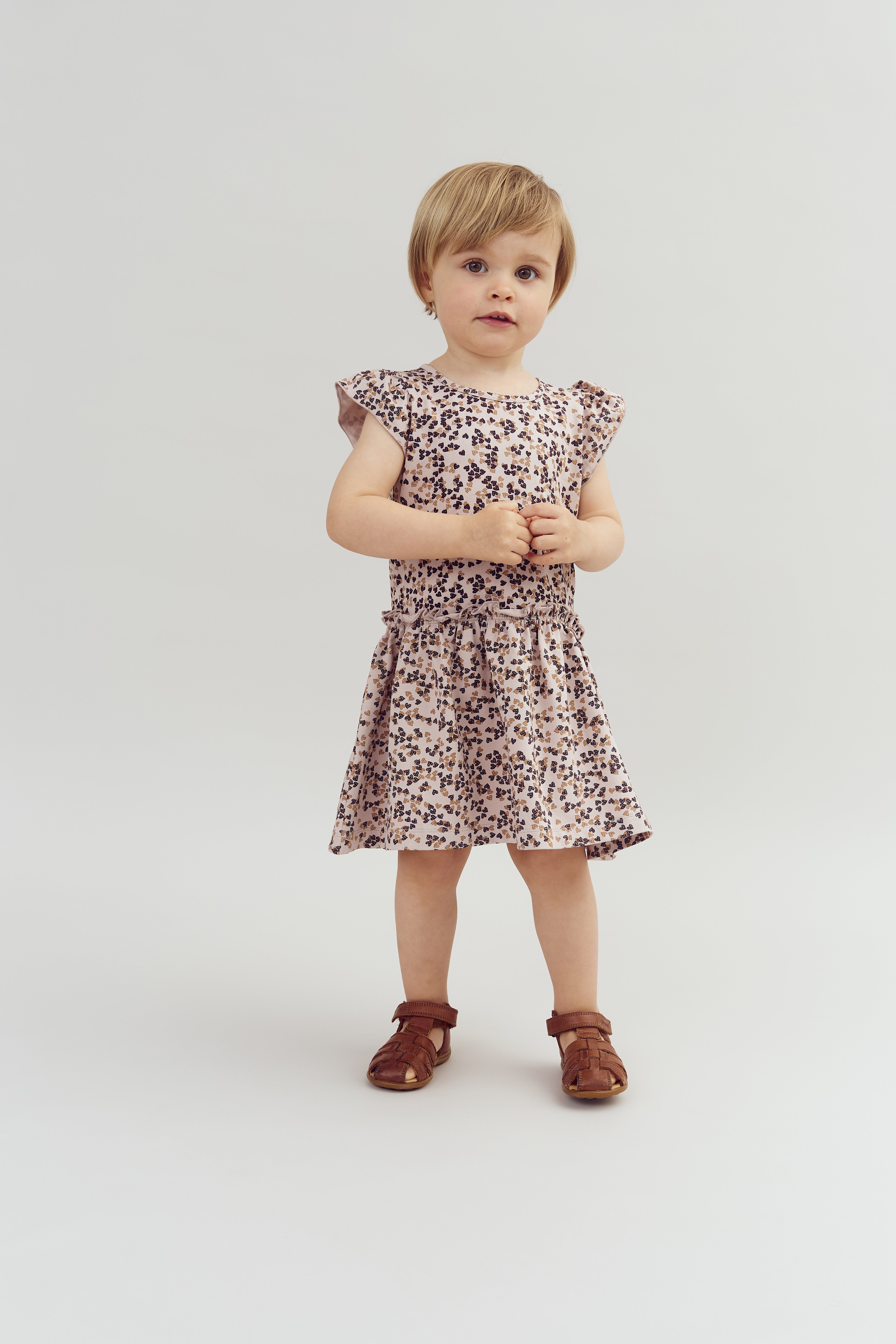 A young child stands wearing a patterned dress and brown shoes, hands clasped. The background is plain, white, and minimalistic.