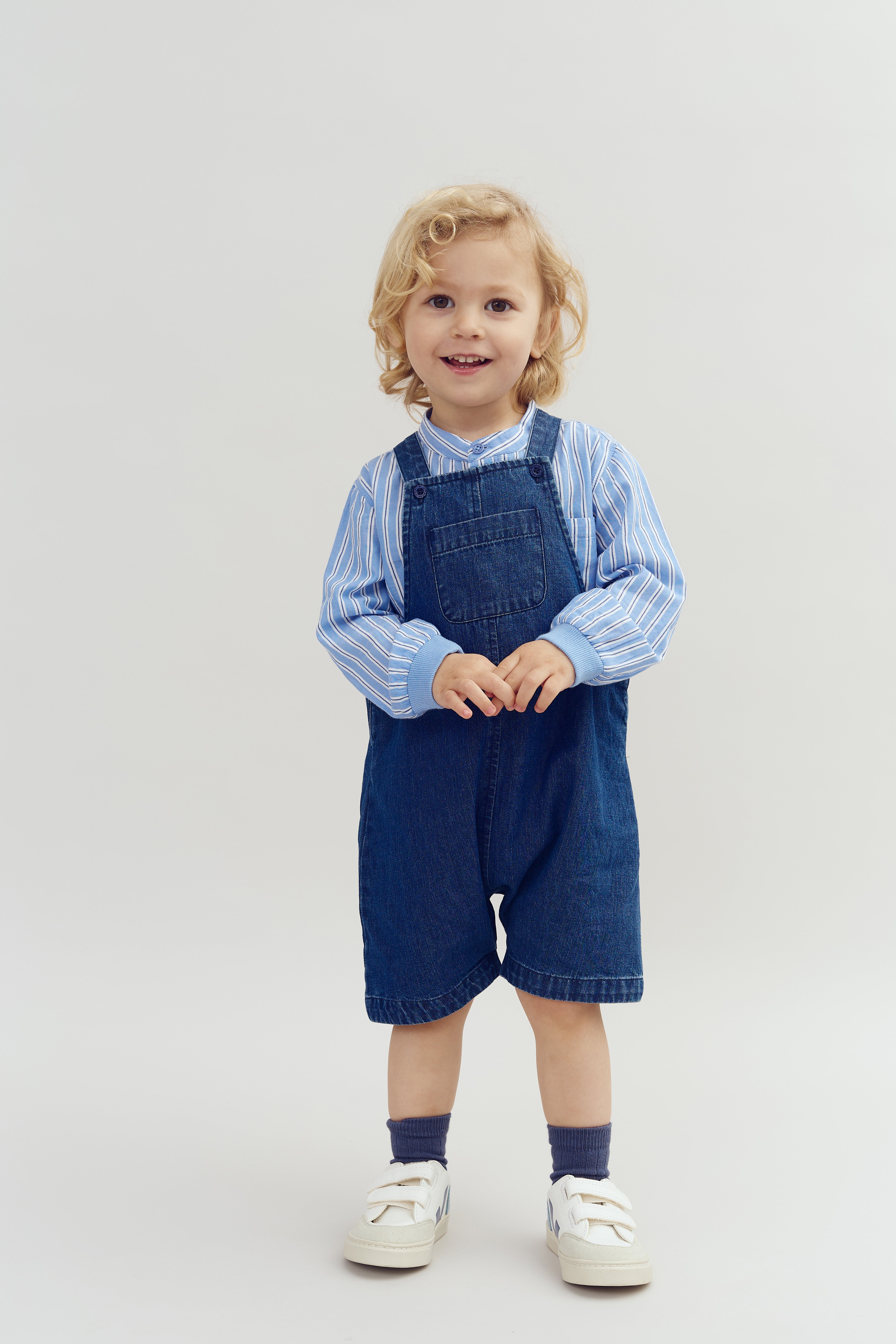 Een jong kind staat lachend in de camera. Het draagt een blauwe gestreepte blouse met denim tuinbroek en witte sneakers. De achtergrond is een effen, lichte kleur.
