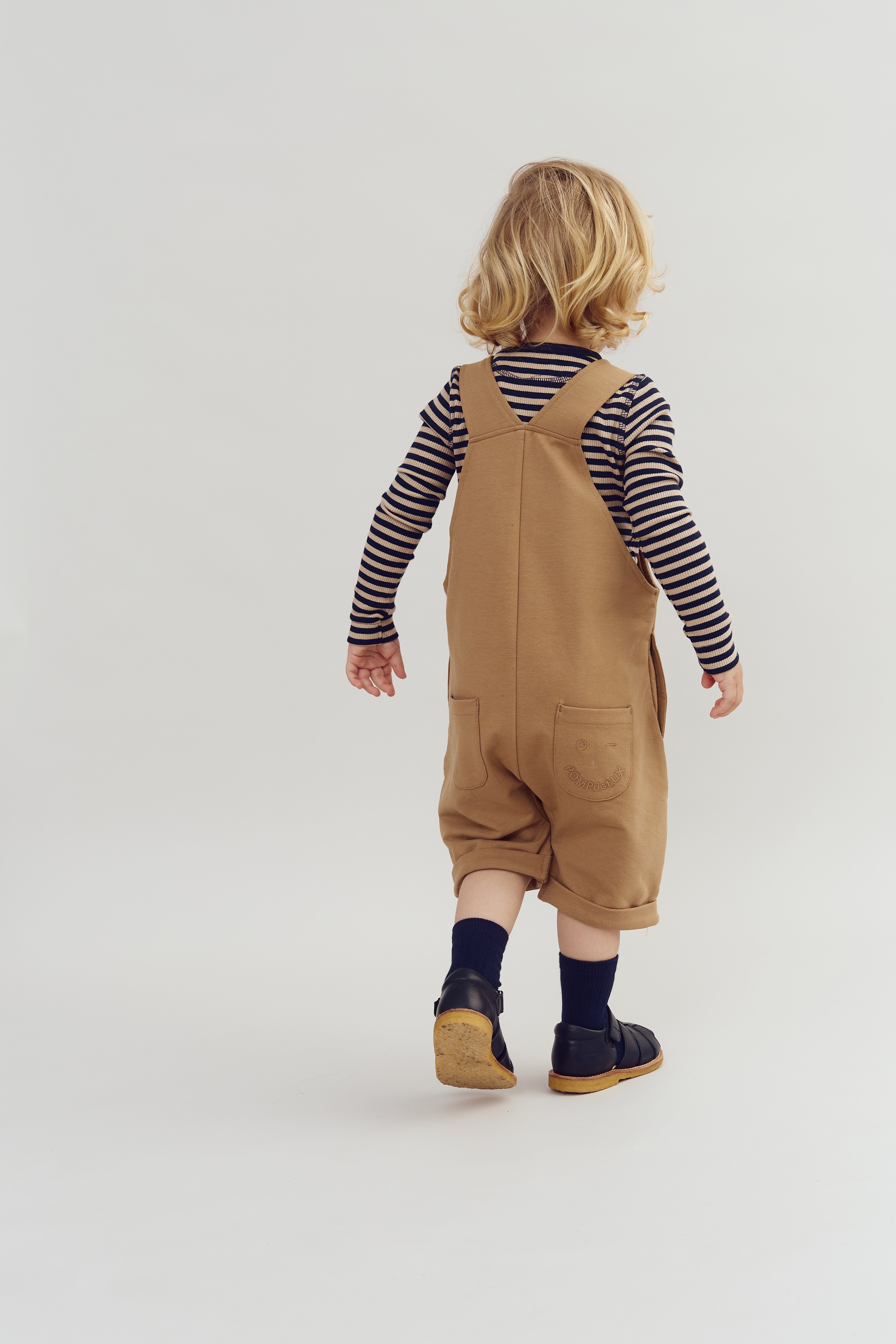A young child wearing a striped shirt and brown overalls walks away on a plain white background, creating a simple and neutral setting.