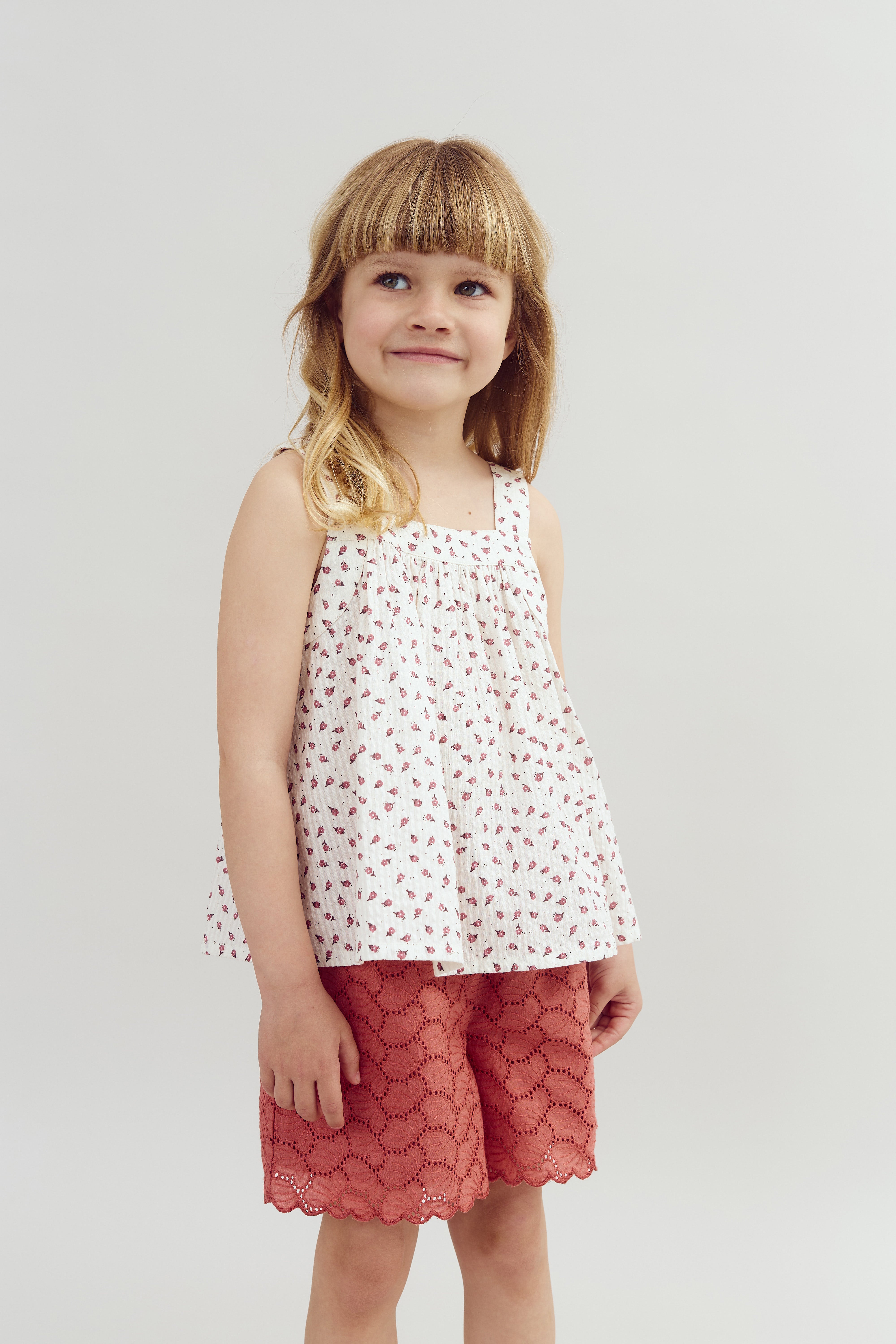 A child stands smiling, wearing a sleeveless white top with a floral pattern and red lace shorts, set against a plain light background.