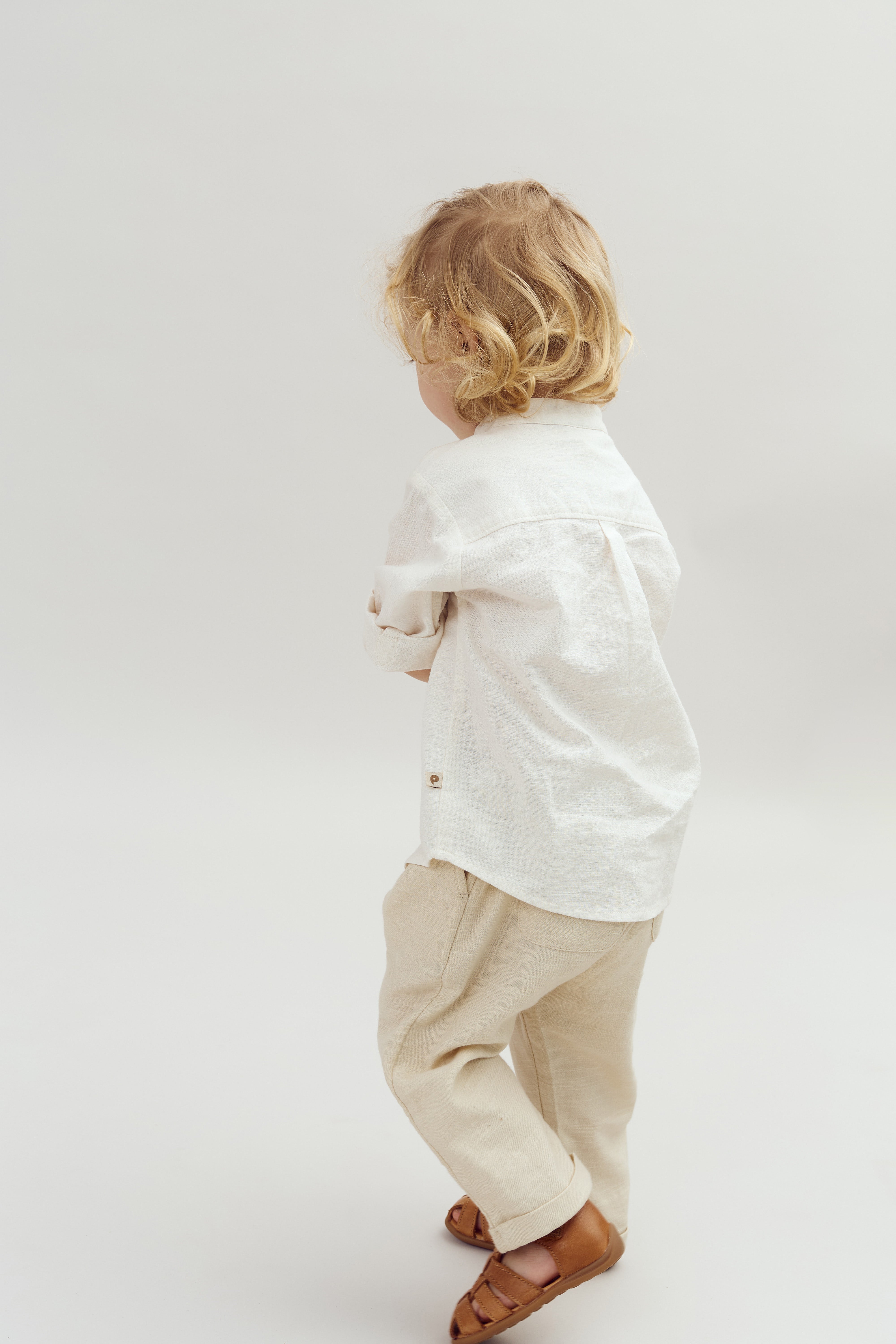 Een klein kind met krullend haar draagt een witte blouse en beige broek, terwijl het op een witte achtergrond staat. Het kindje heeft bruine sandalen aan.