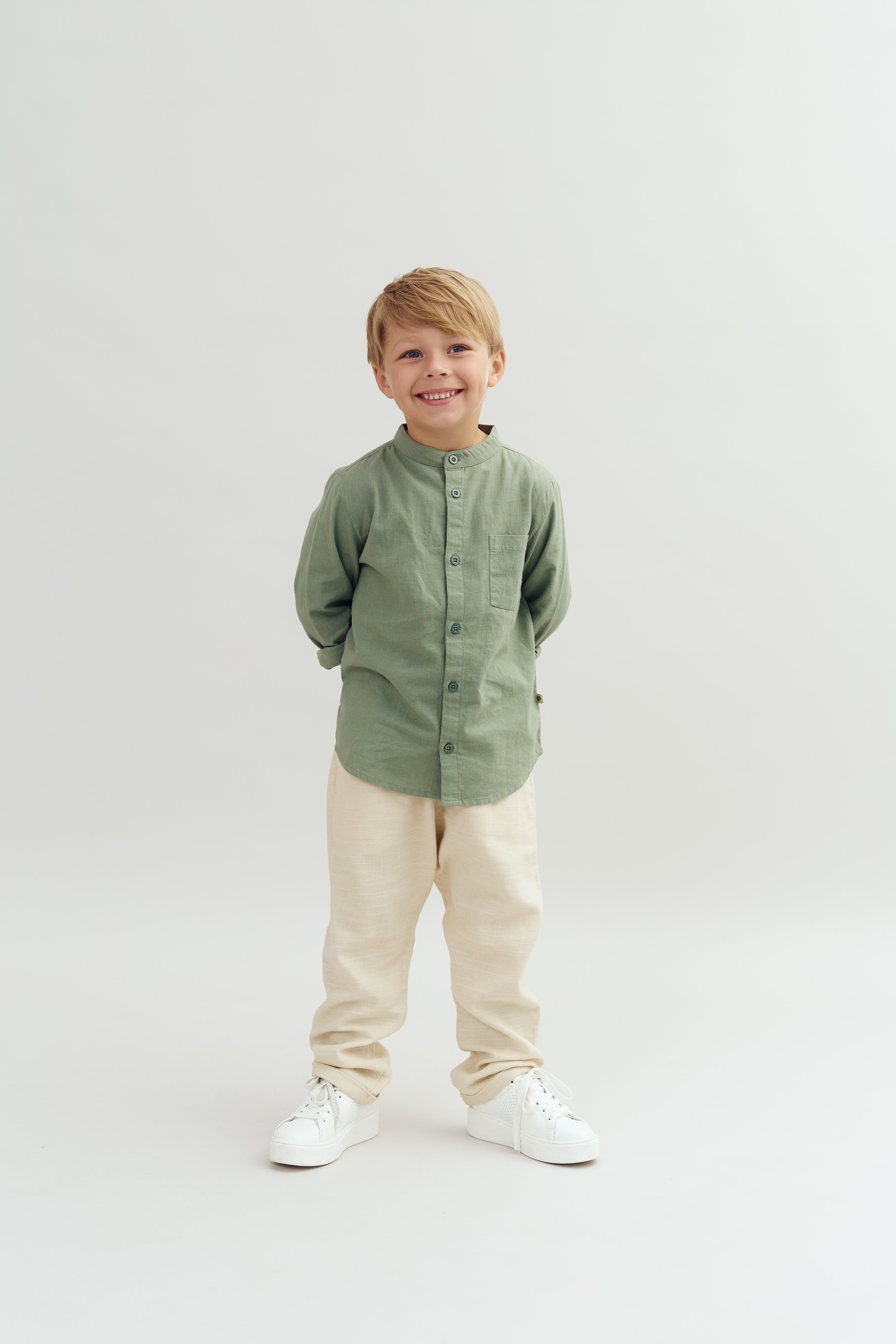 Een jong kind staat glimlachend, gekleed in een groene blouse en beige broek, met handen achter de rug in een witte, minimalistische ruimte.