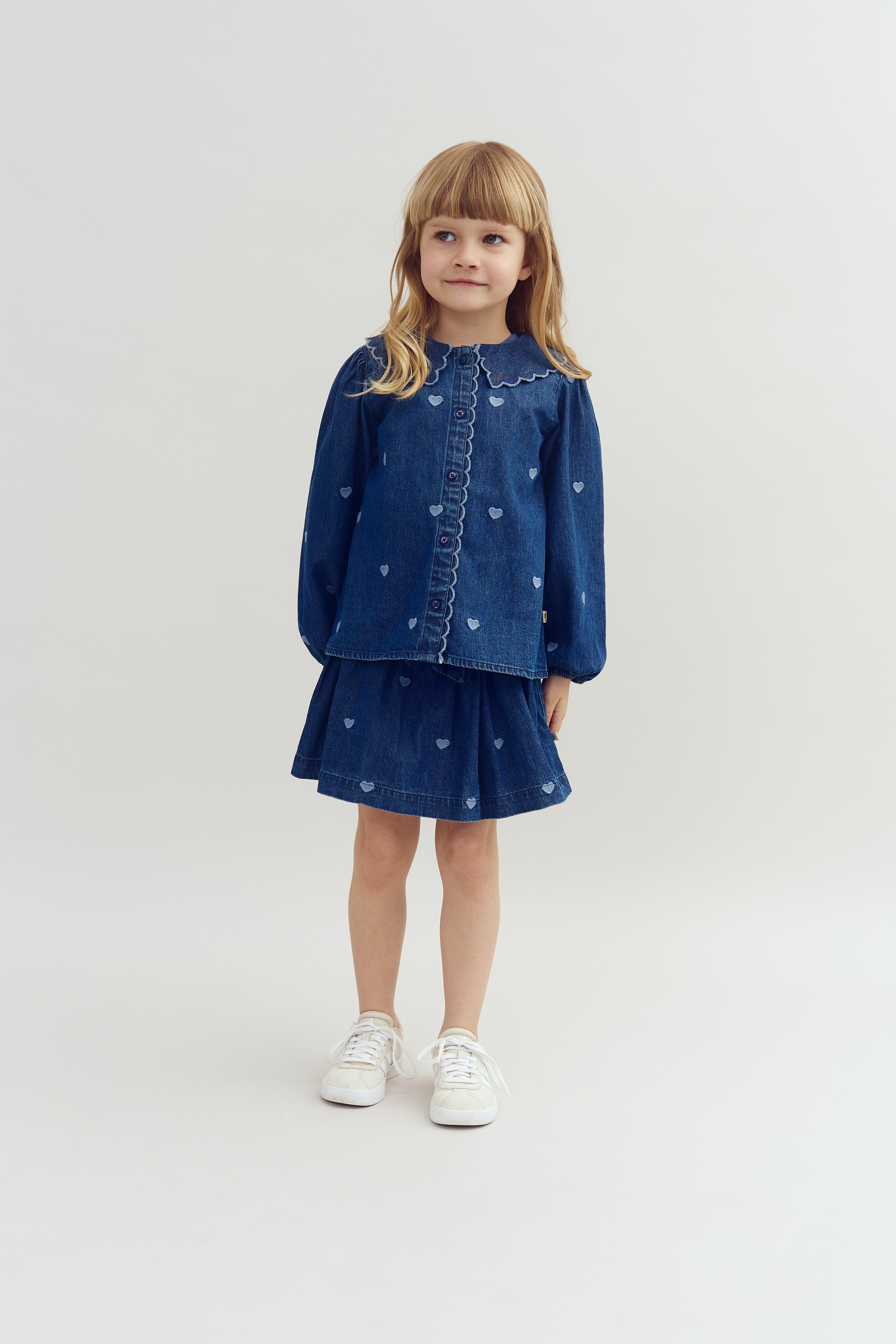 Een jong meisje poseert in een denim outfit met een hartjespatroon, bestaande uit een blouse en rok. Ze staat in een eenvoudige, lichte ruimte en draagt witte sneakers.