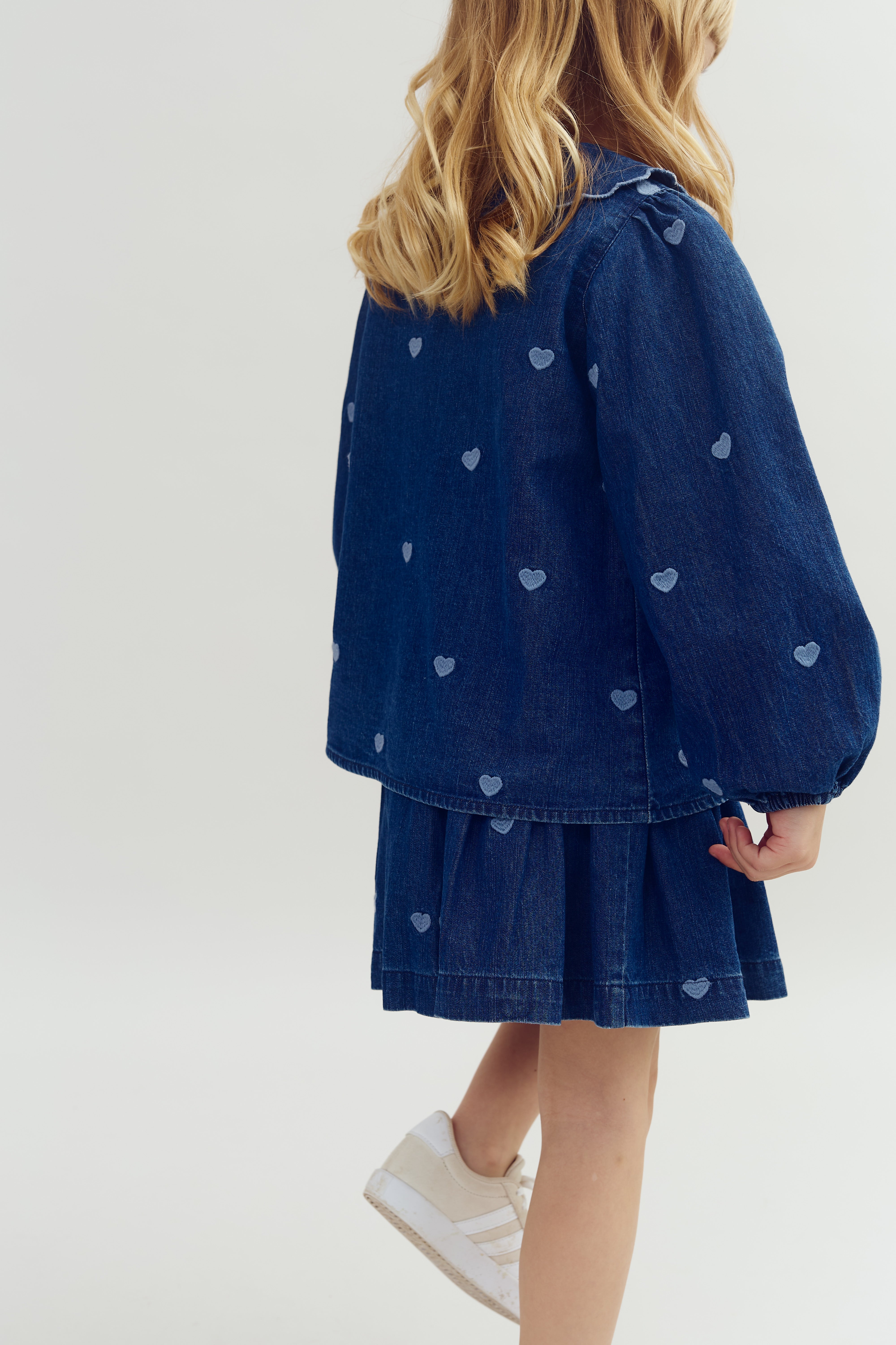 Een jong kind draagt een blauw spijkerrokje en blouse met hartpatronen, staand op een witte achtergrond, met witte sportschoenen. Het haar is blond en golvend.
