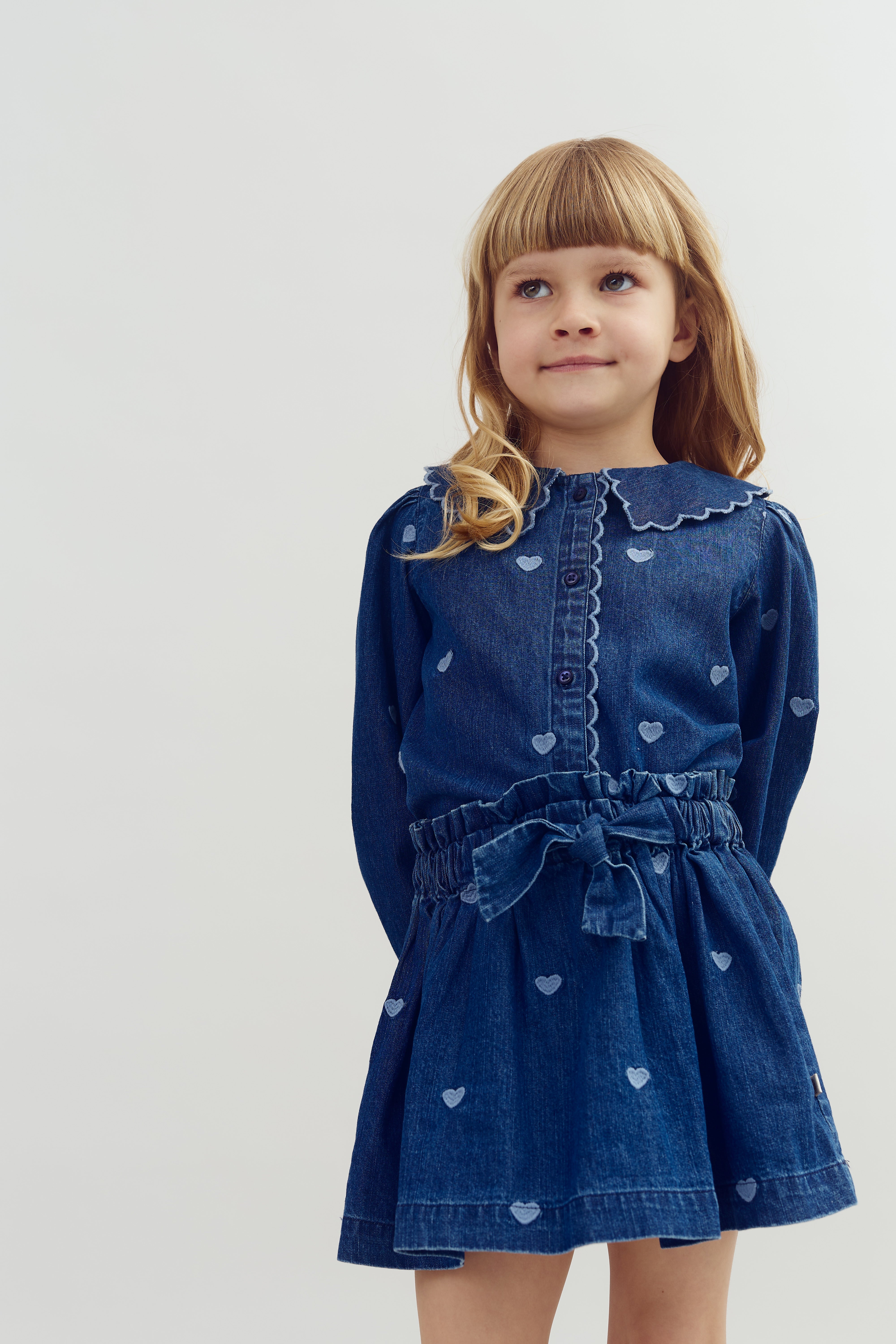 Meisje in blauwe jurk met hartjes, kijkt omhoog met een lichte glimlach, tegen een effen, witte achtergrond.