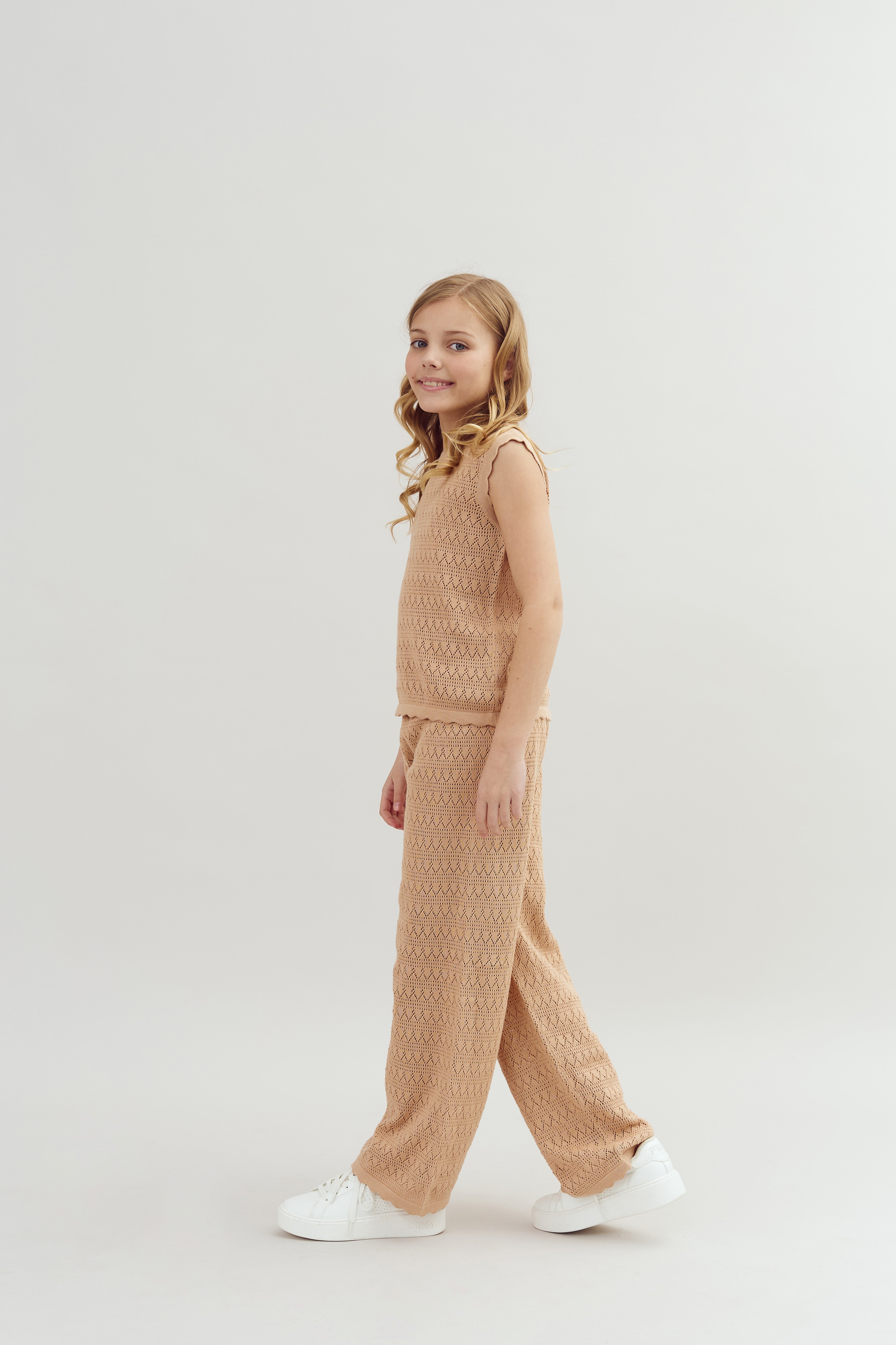 A young girl stands, wearing a tan knitted sleeveless top and matching pants with white sneakers, in a simple, neutral studio setting.