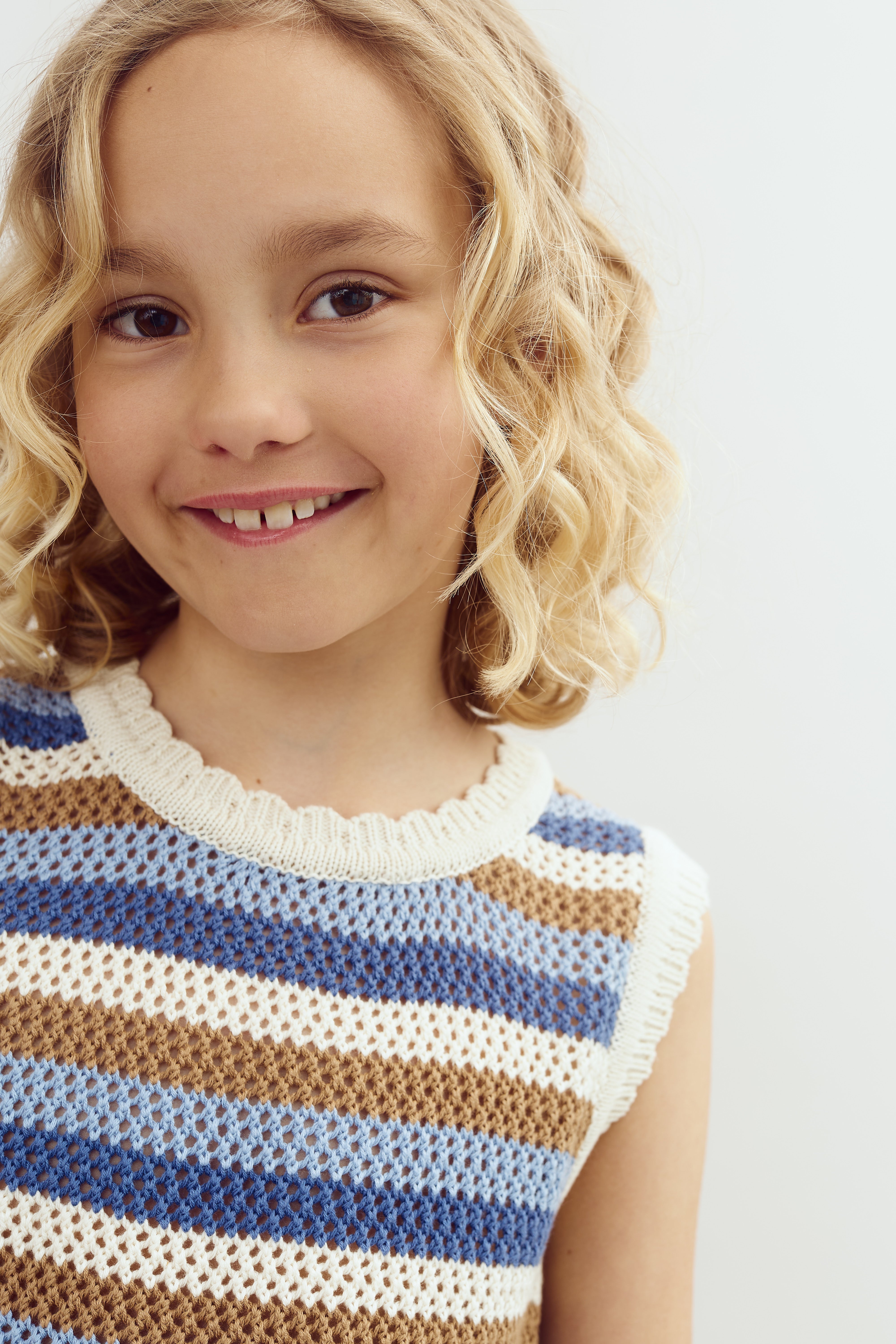 A young girl with curly blonde hair smiles, wearing a striped, crocheted top in blue, brown, and white. She is in front of a plain, light-colored background.