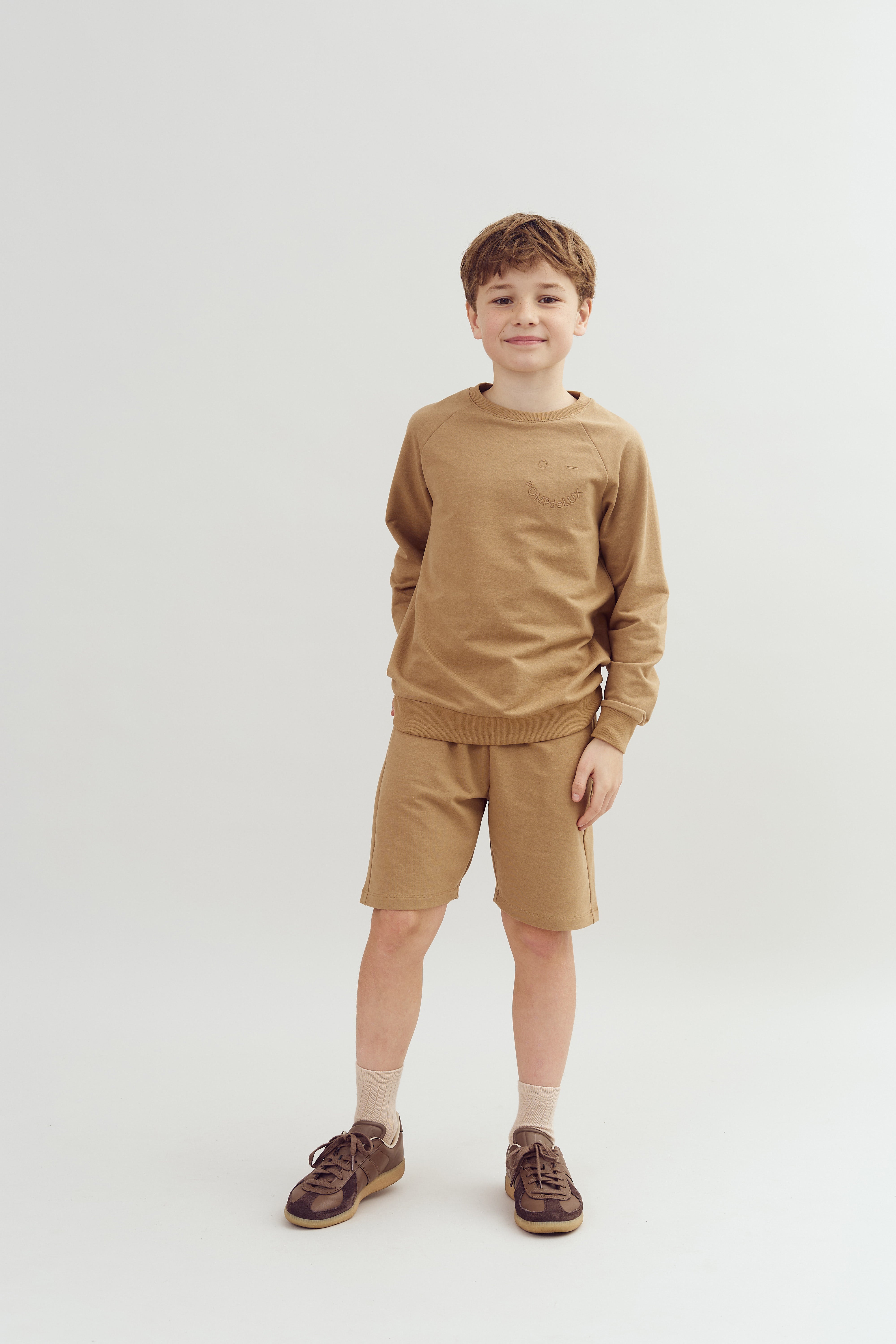 A child stands smiling, wearing a brown sweatshirt and shorts with dark sneakers and light socks, in a plain white studio setting.