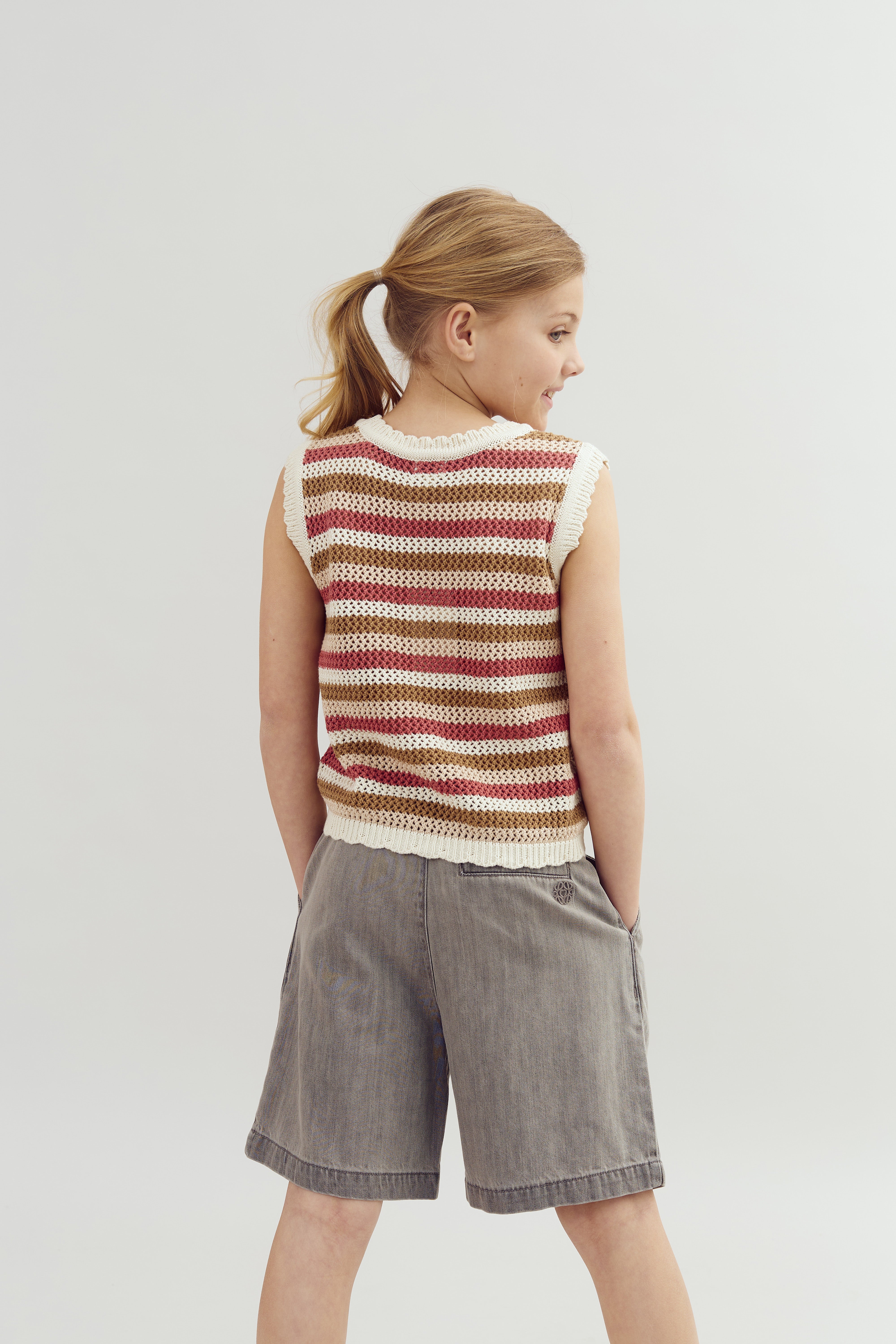 Een kind draagt een gestreepte gebreide trui, staand met de rug naar de camera. Grijze korte broek, lichte achtergrond, haar in een staart.