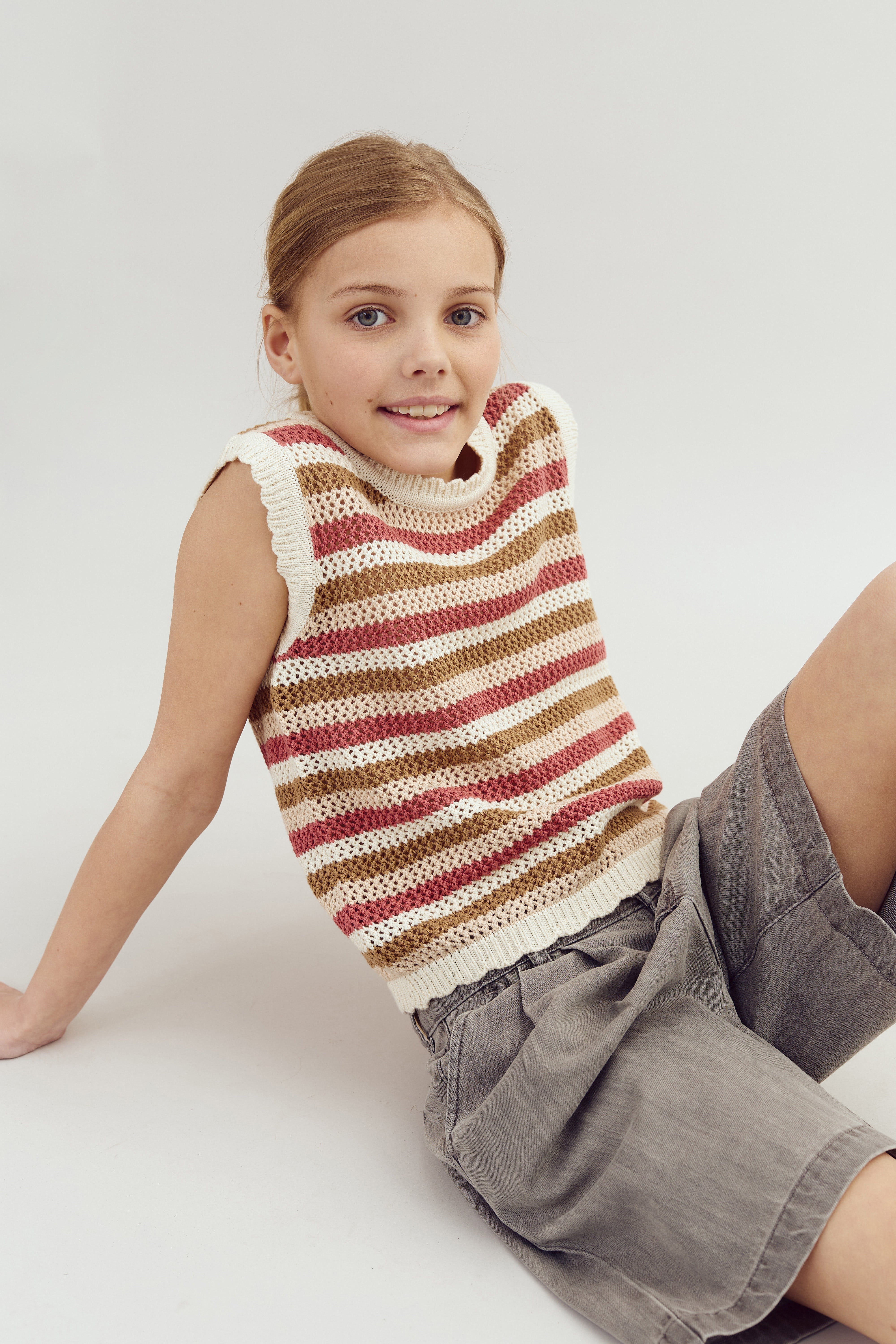 Child sitting on the floor, wearing a striped vest with red, beige, and white colors and gray shorts, against a neutral background.