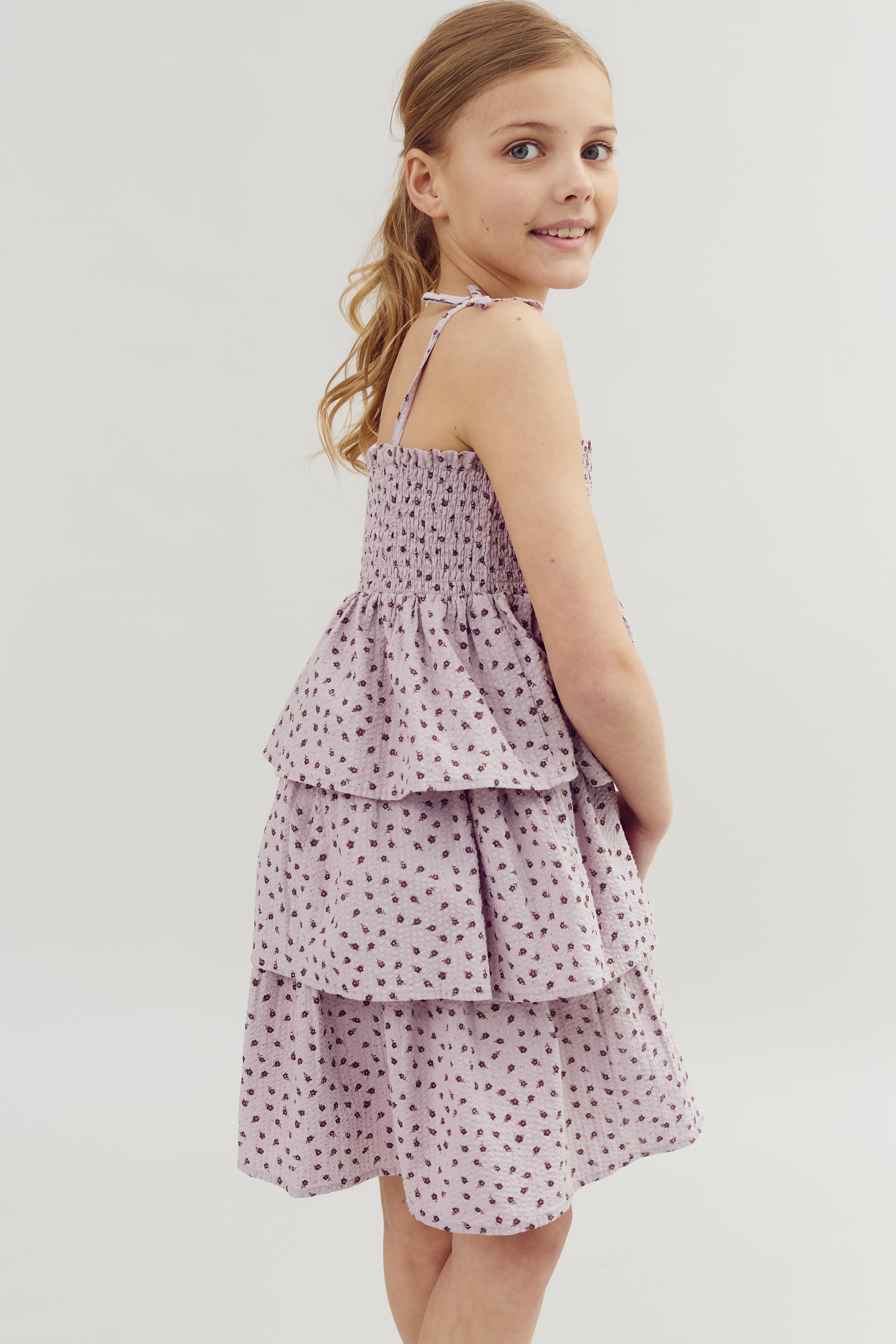 A girl stands smiling, wearing a sleeveless, lilac tiered dress with a floral pattern, in a neutral, light-colored setting.
