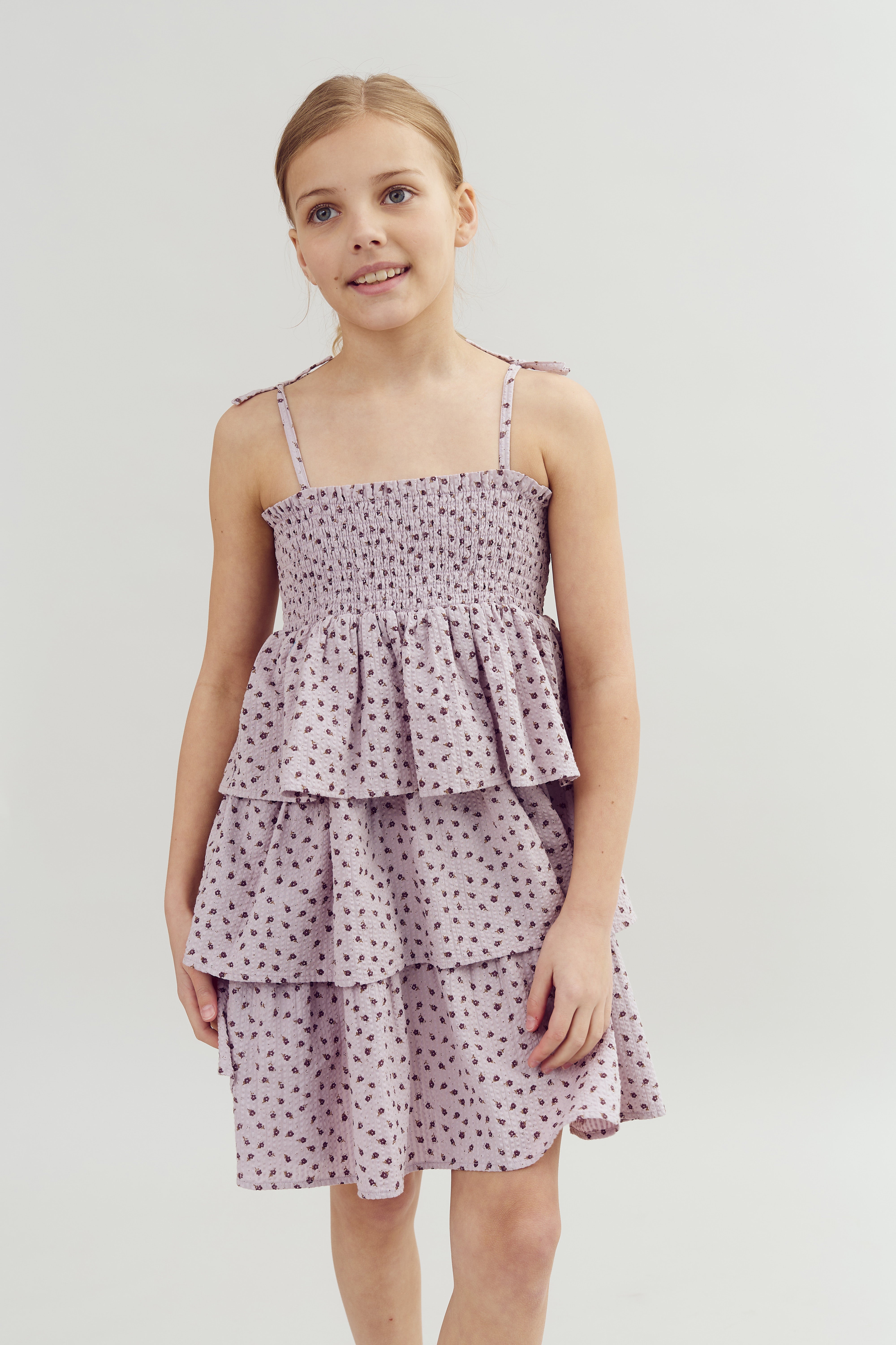 A smiling child stands wearing a tiered, sleeveless lavender dress with a small print, set against a plain light background.