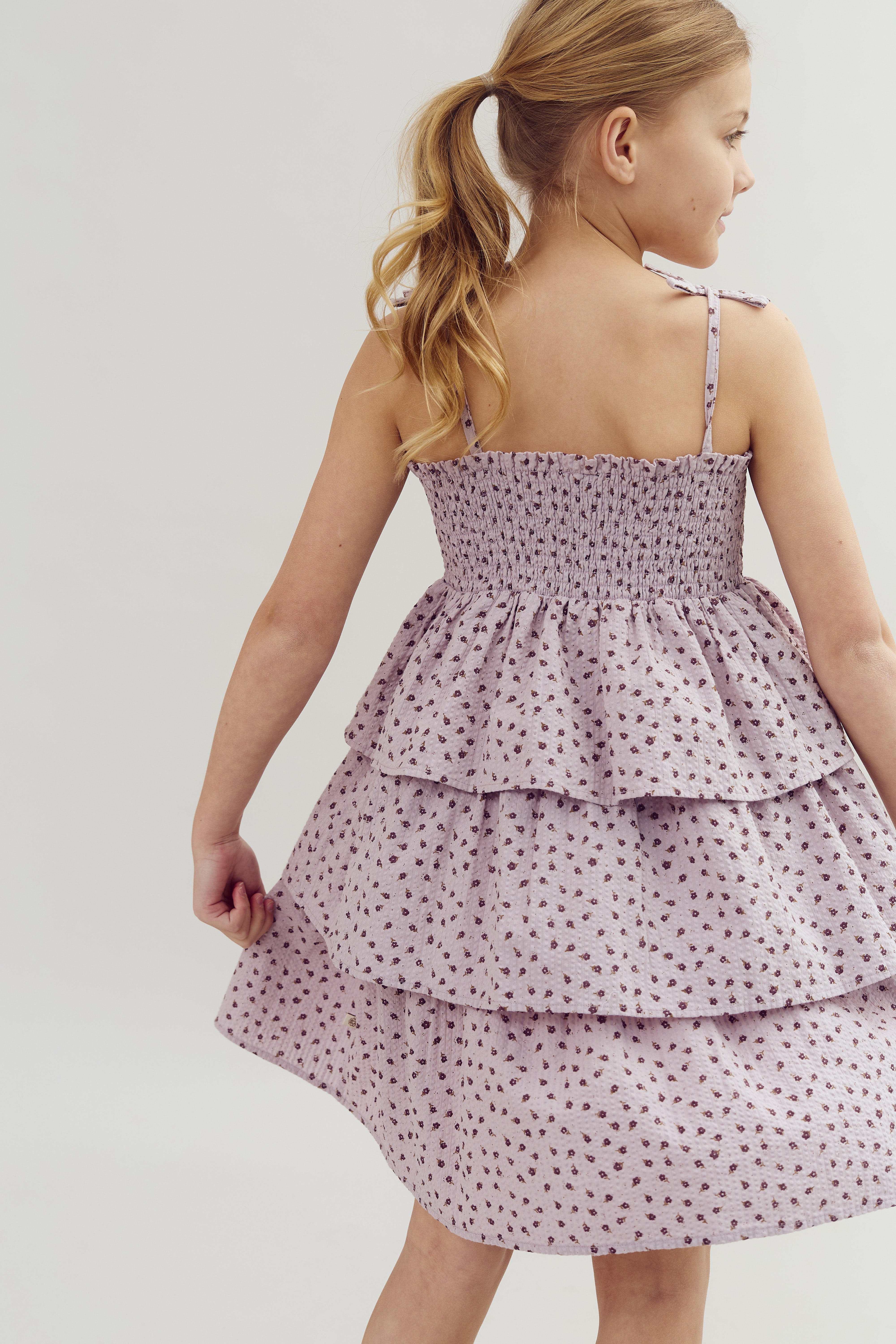 A child, with blonde hair in a ponytail, wears a light purple, tiered dress with small floral patterns, standing against a plain, light background.