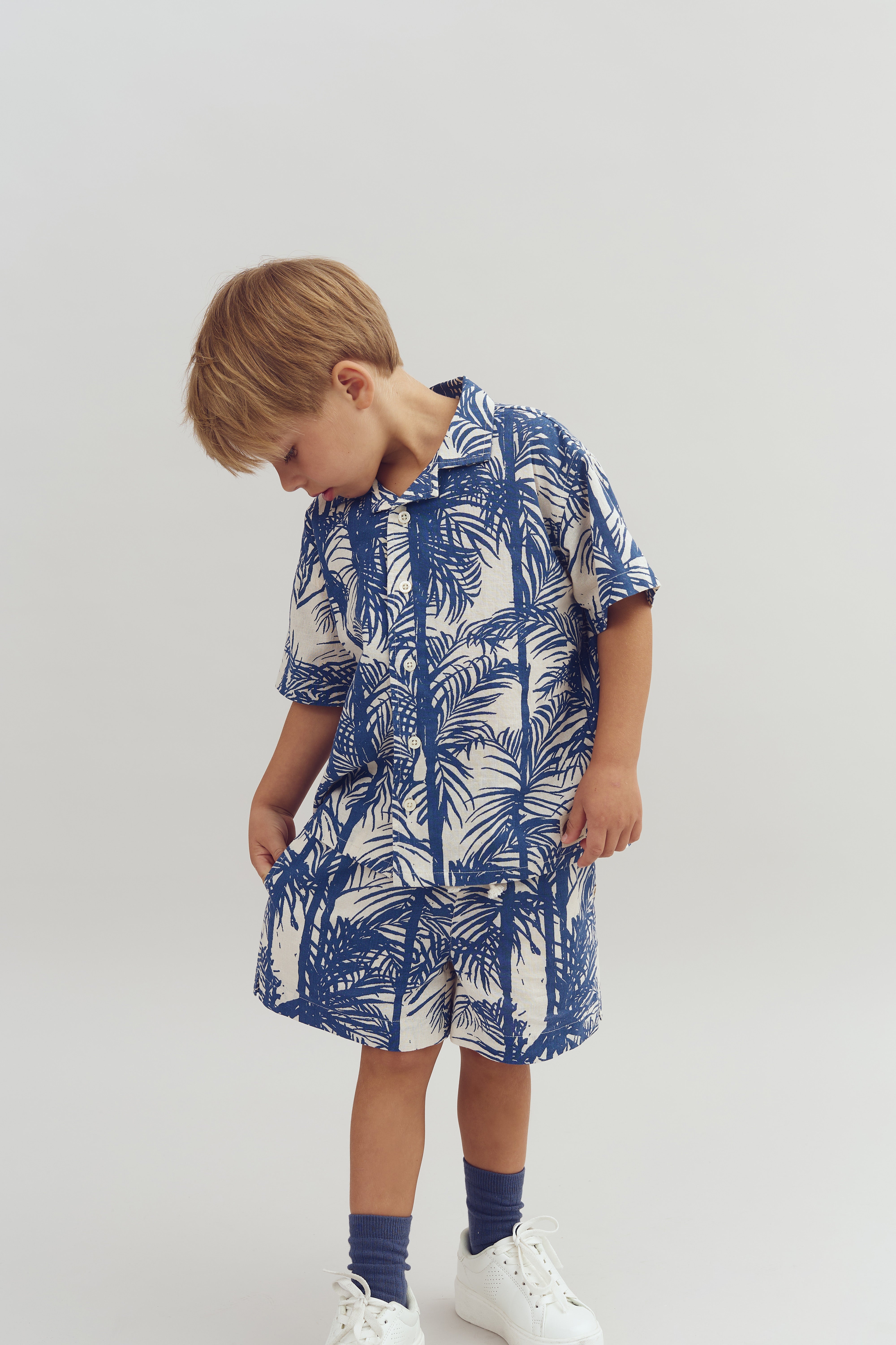 Een kind draagt een blauw-wit palmboompatroon kleding, kijkt omlaag en heeft zijn hand in de zakken. Hij staat in een neutrale, lichte omgeving.