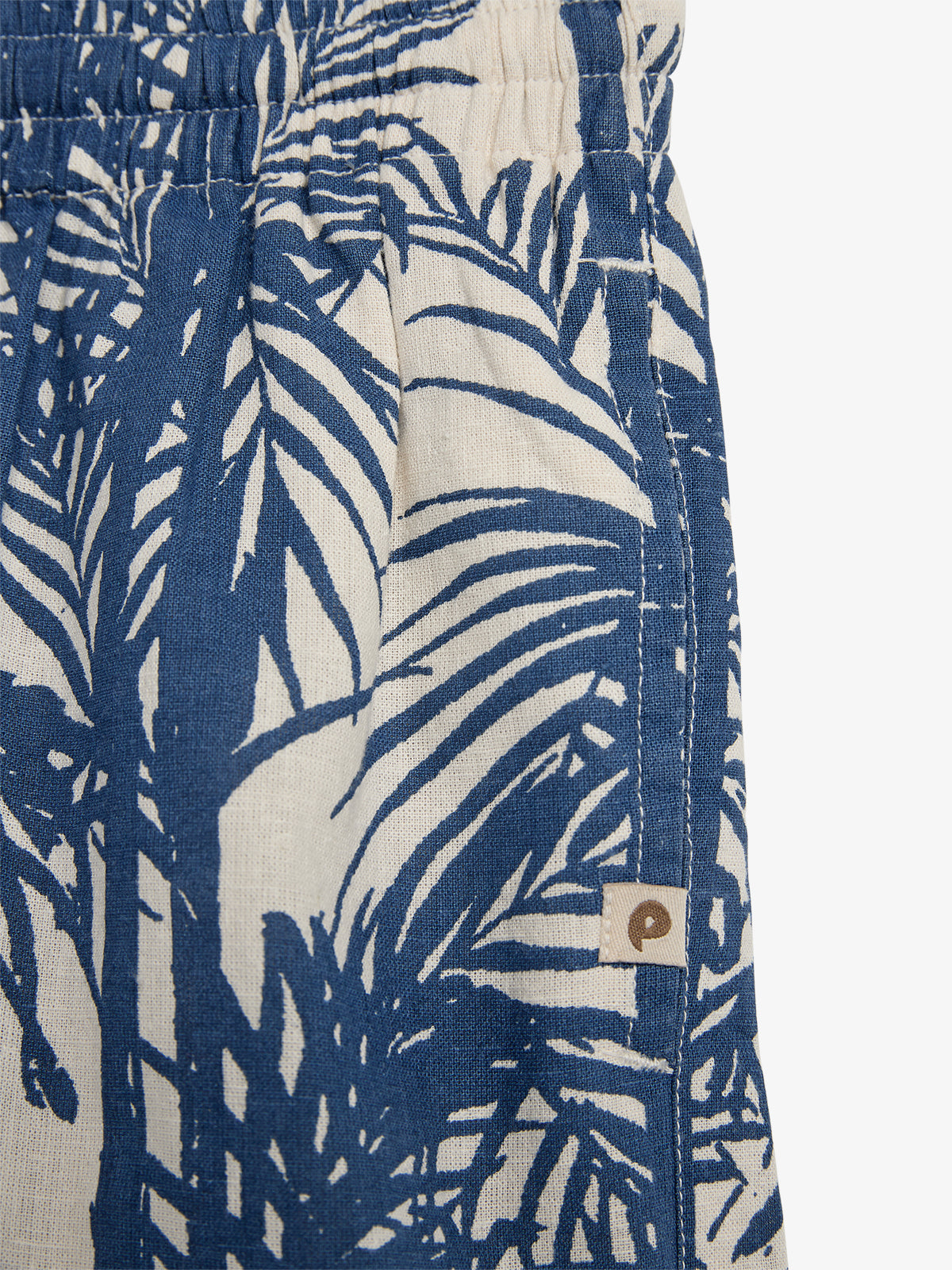 Een stuk stof met palmbladeren in blauw-wit patroon, gedeeltelijk zichtbaar. Het heeft een elastische tailleband. Er is een klein label met een bruin logo erop genaaid.
