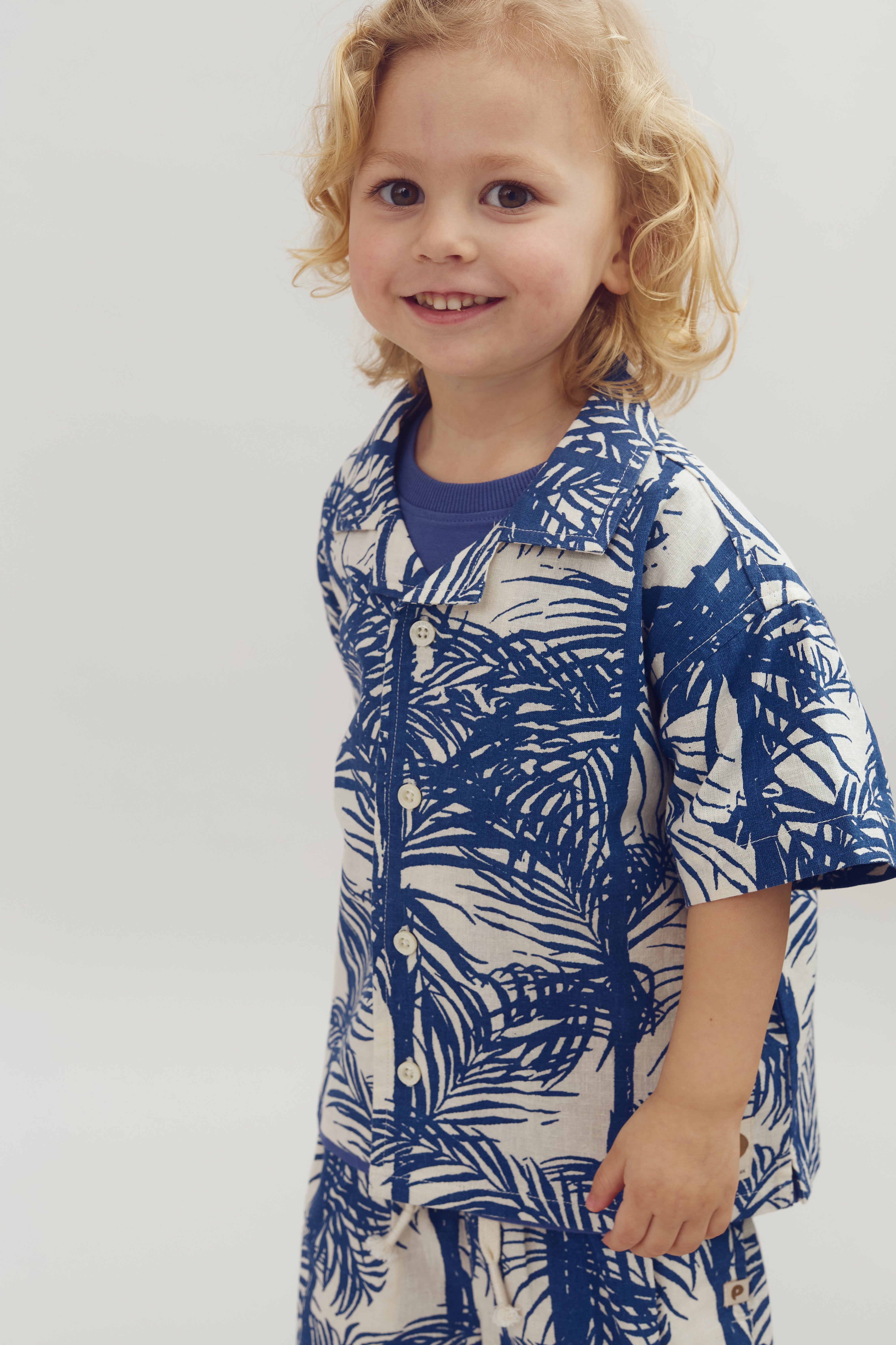Kind met blond haar draagt een blauw-wit shirt met bladerenmotief. Hij lacht en staat in een eenvoudige, lichte ruimte.