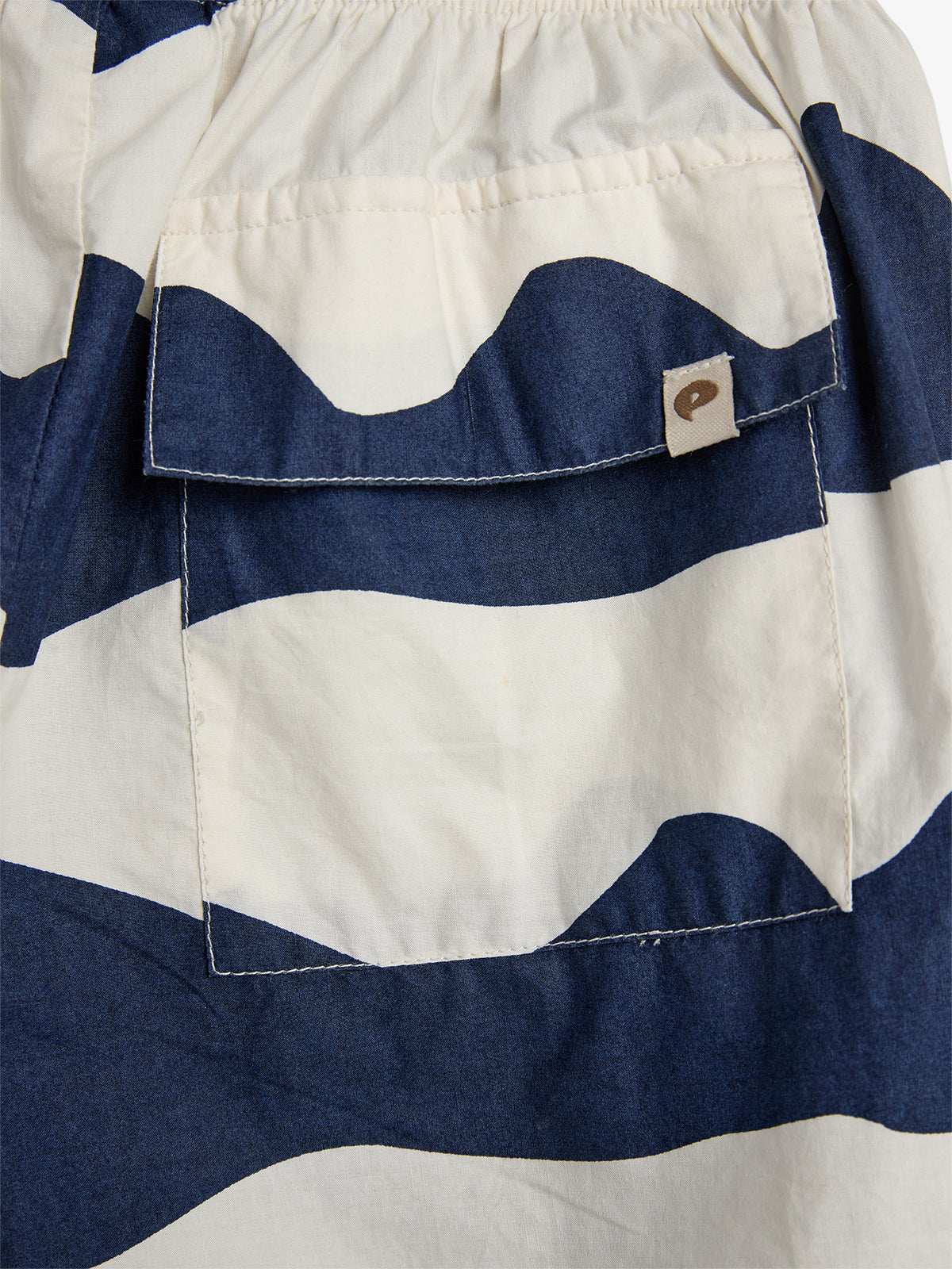 Een kledingstuk met blauw-witte golvende strepen en een zak met flap, waarop een label met een logo is bevestigd. Alles bevindt zich op een lichtgekleurde achtergrond.