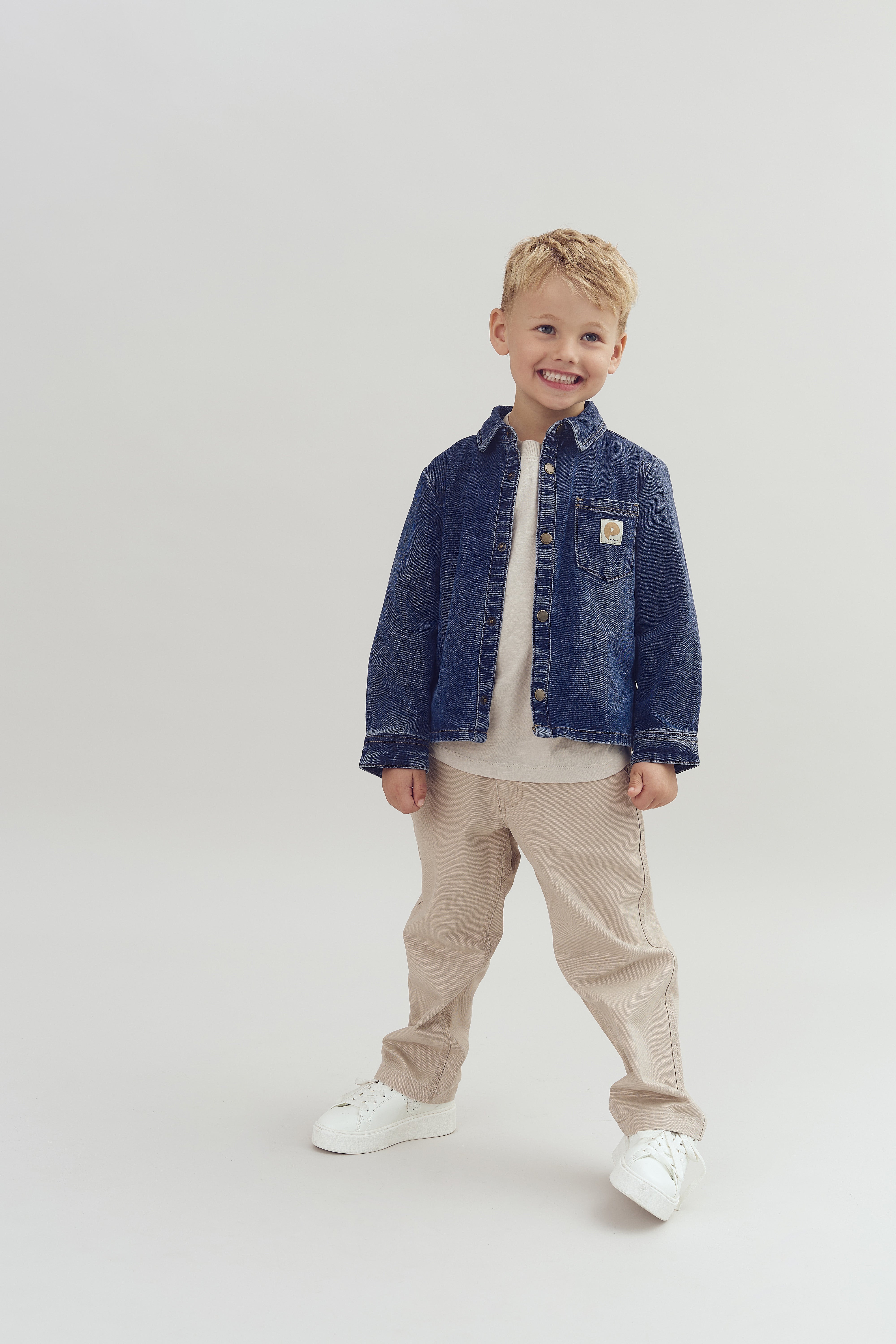 Een jong kind lacht, gekleed in een blauw spijkerjack, beige broek en witte schoenen, staand op een neutrale achtergrond.