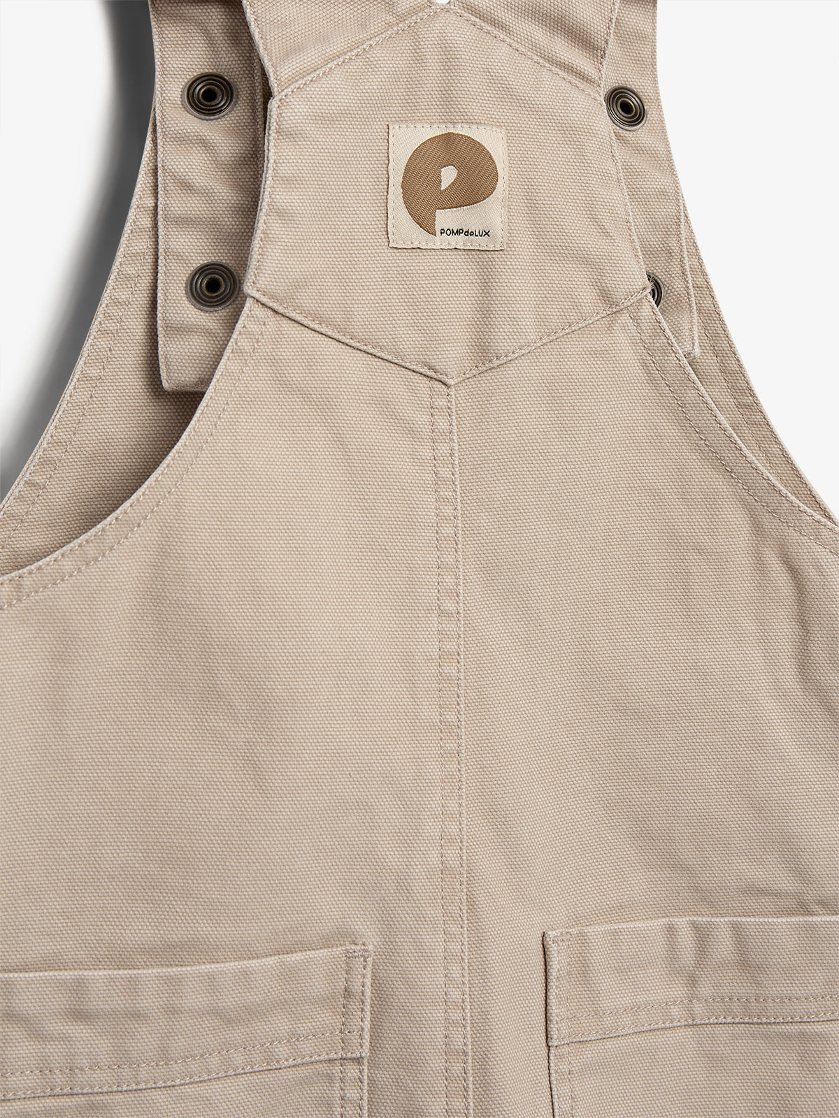 Beige fabric overalls with buttoned straps and a logo patch reading "POMPdeLUX" attached on the chest. The garment features stitched pockets and is designed for a practical and stylish look.