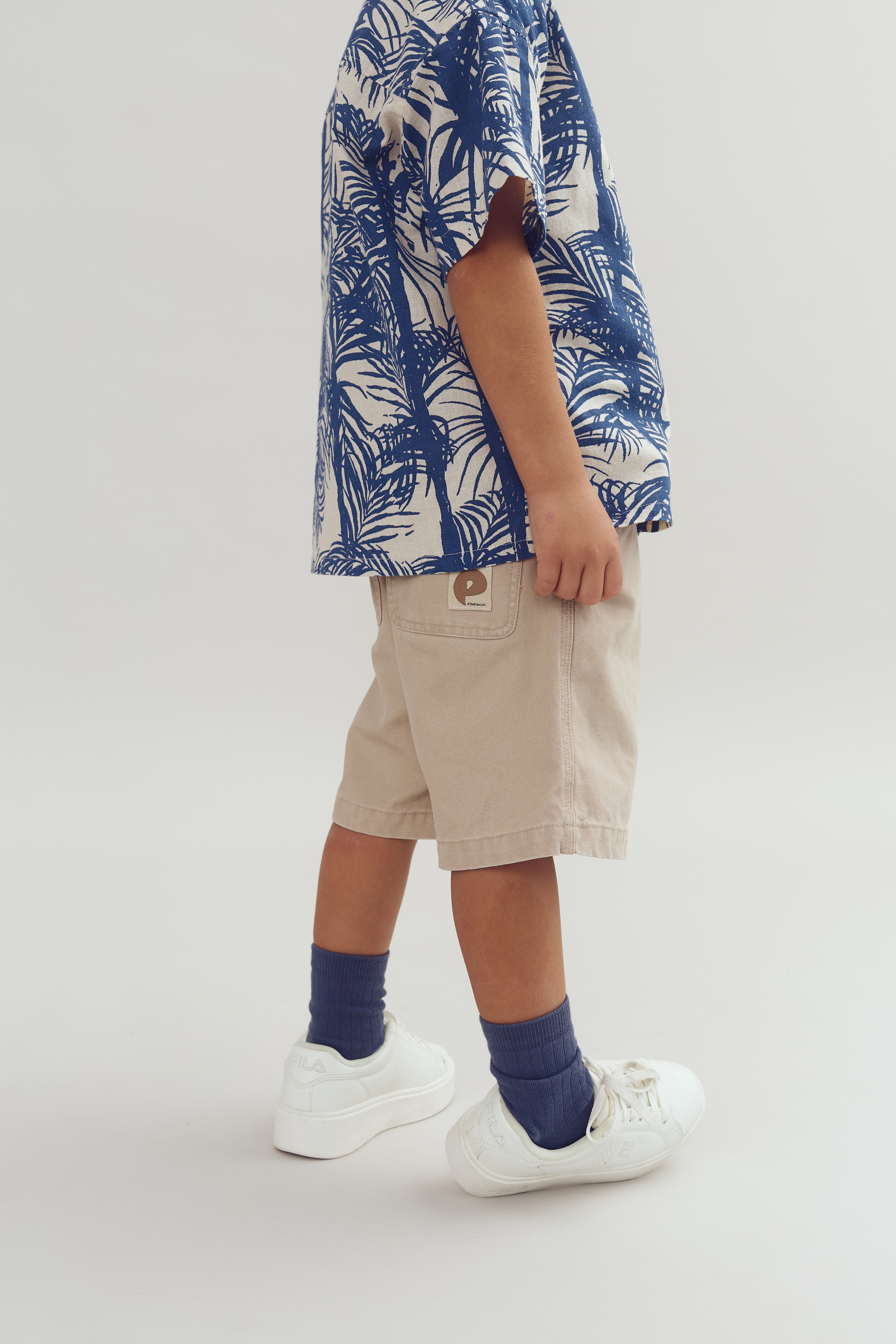 Kind met beige korte broek en blauw-wit overhemd met bladmotief, draagt witte Fila sneakers en blauwe sokken, staand tegen een witte achtergrond.