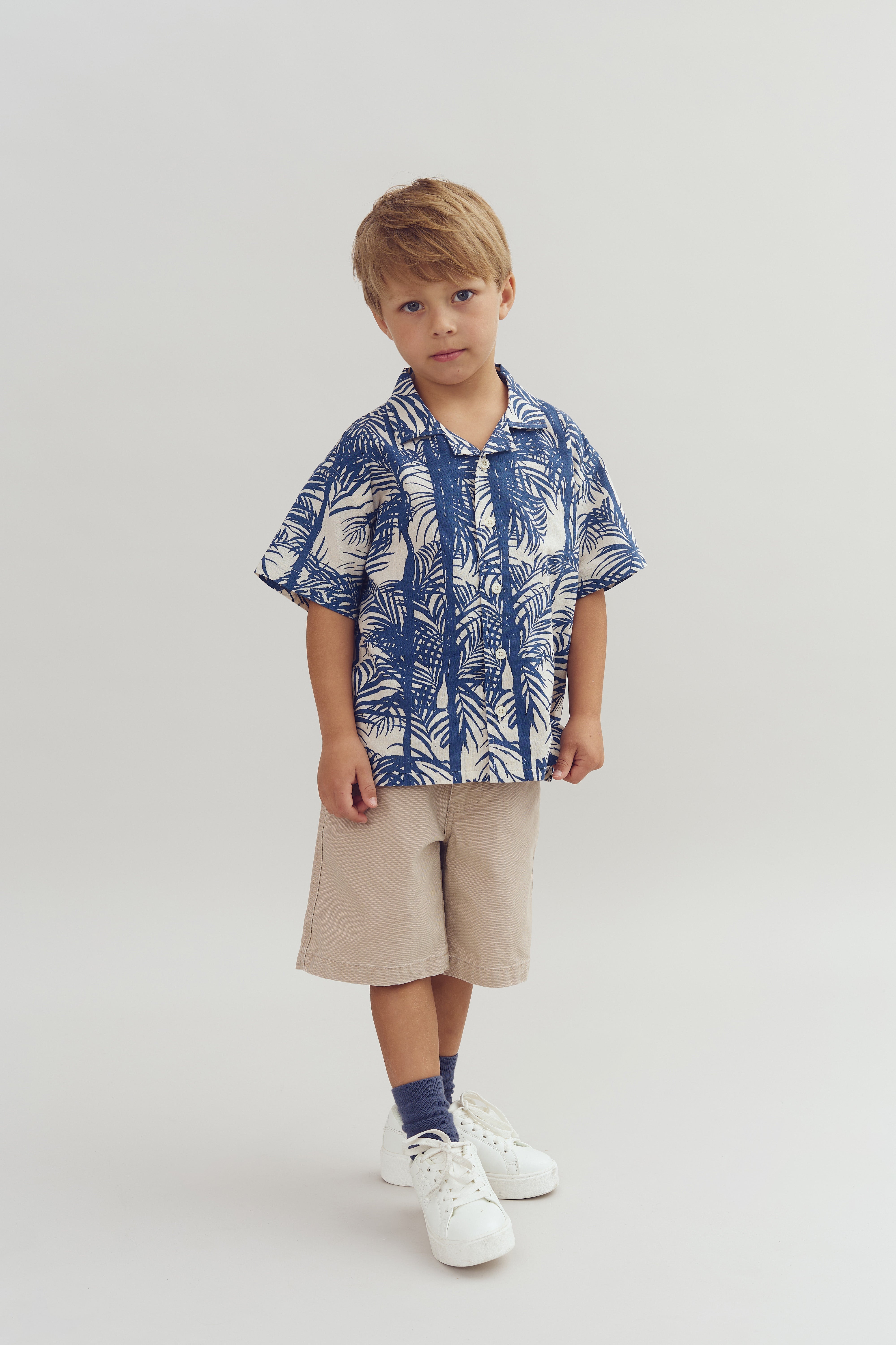 Een jongetje draagt een blauw-wit gebloemd overhemd met beige korte broek en witte schoenen. Hij staat stil tegen een effen, lichte achtergrond.