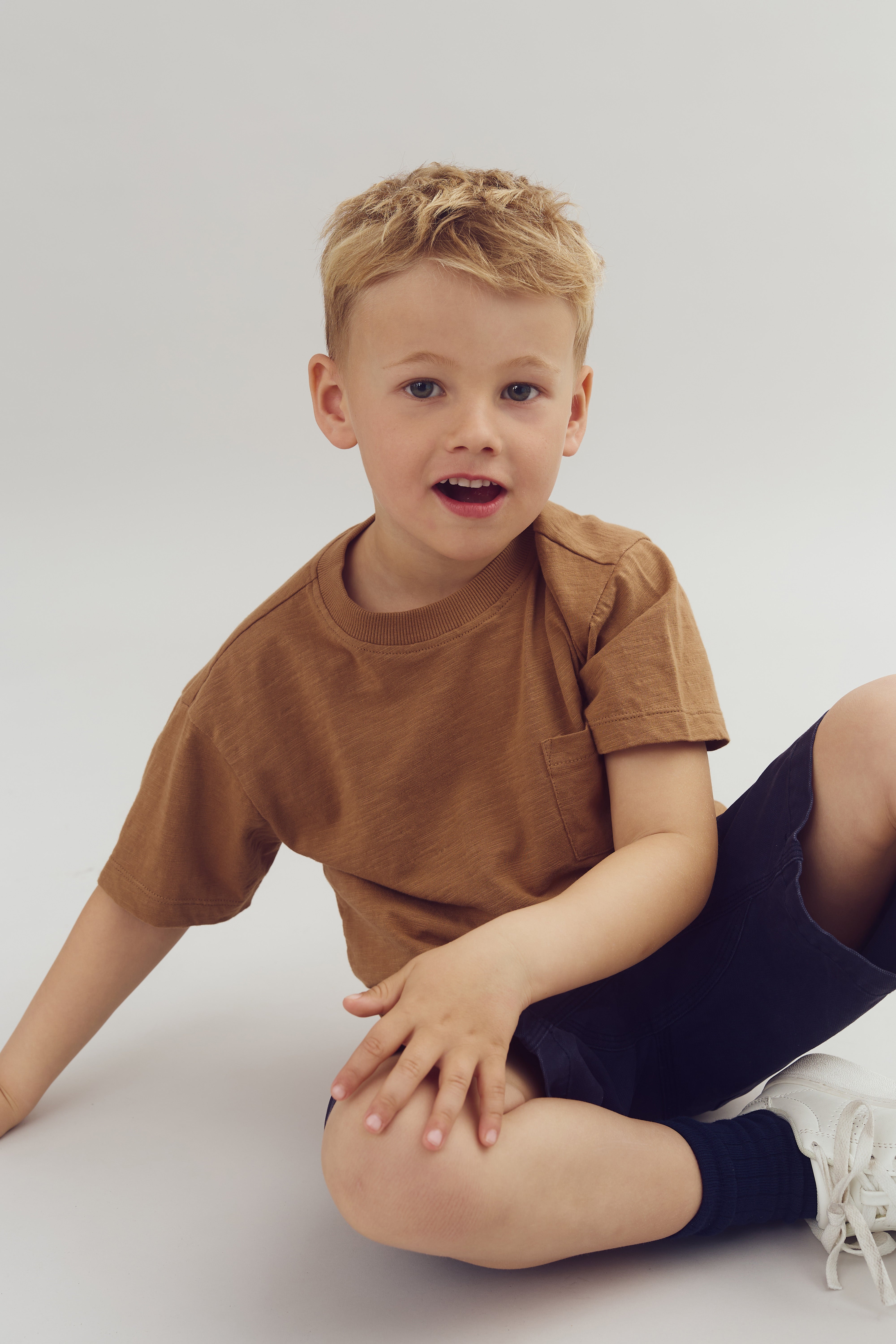 Een jongen zit op de grond, draagt een bruin shirt en donkere shorts. Hij kijkt met een lachende uitdrukking naar de camera in een lichte, neutrale omgeving.