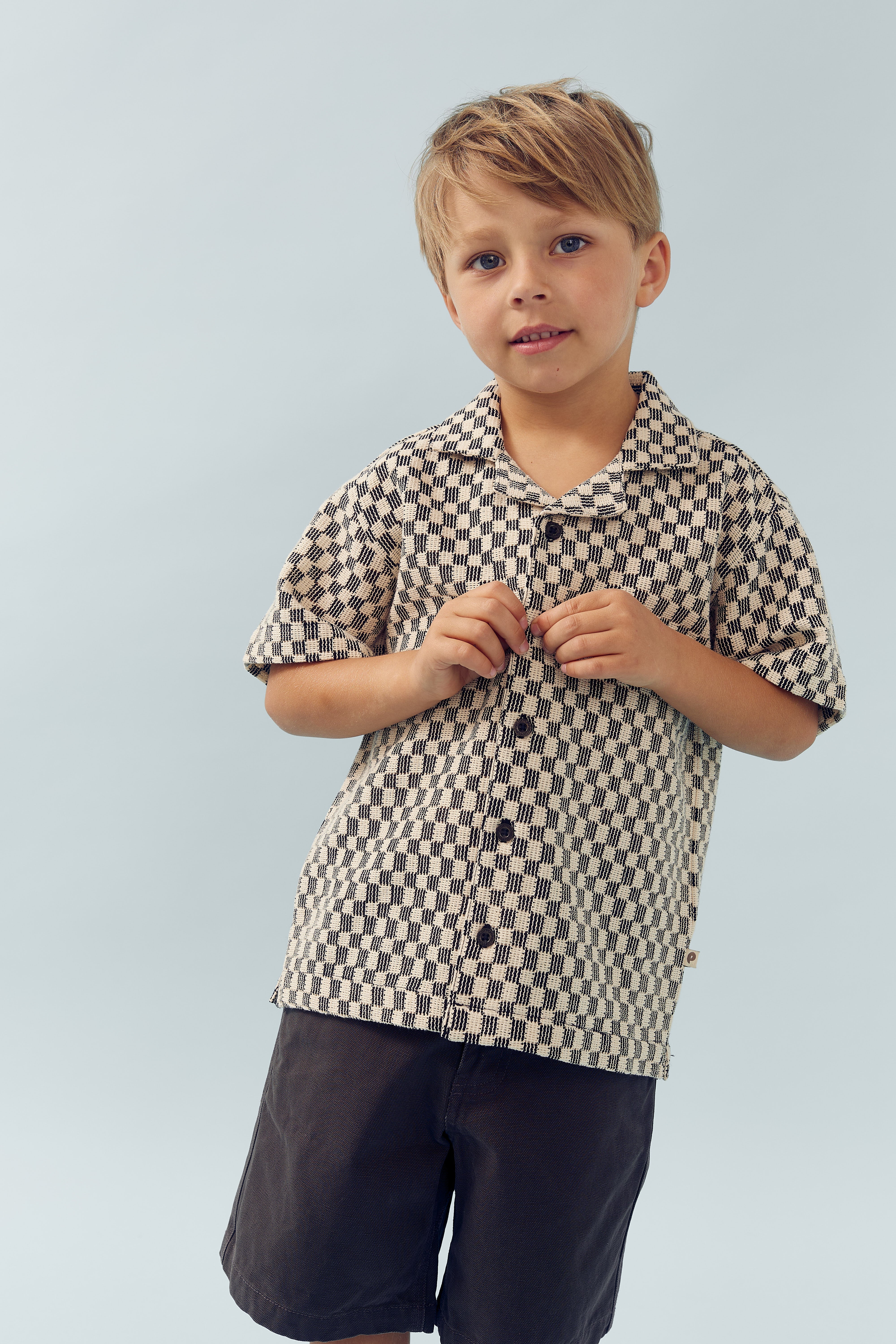 Kind draagt geruite blouse, poseert ontspannen, houdt knoop vast. Neutrale achtergrond benadrukt outfit.