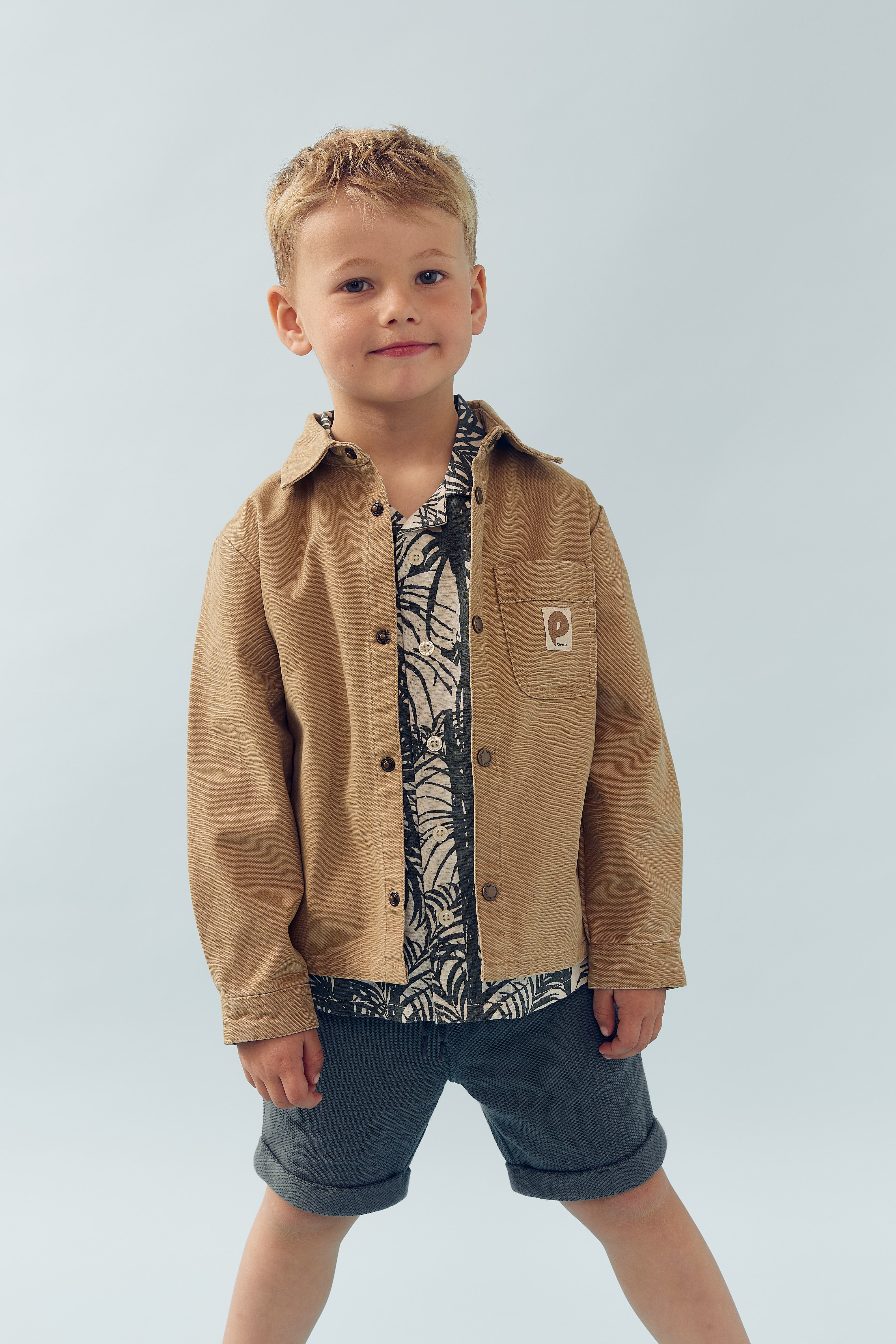 Een jongen draagt een bruin jasje over een shirt met patroon, handen ontspannen langs zijn lichaam, tegen een effen lichtblauwe achtergrond. In de borstzak zit een embleem.