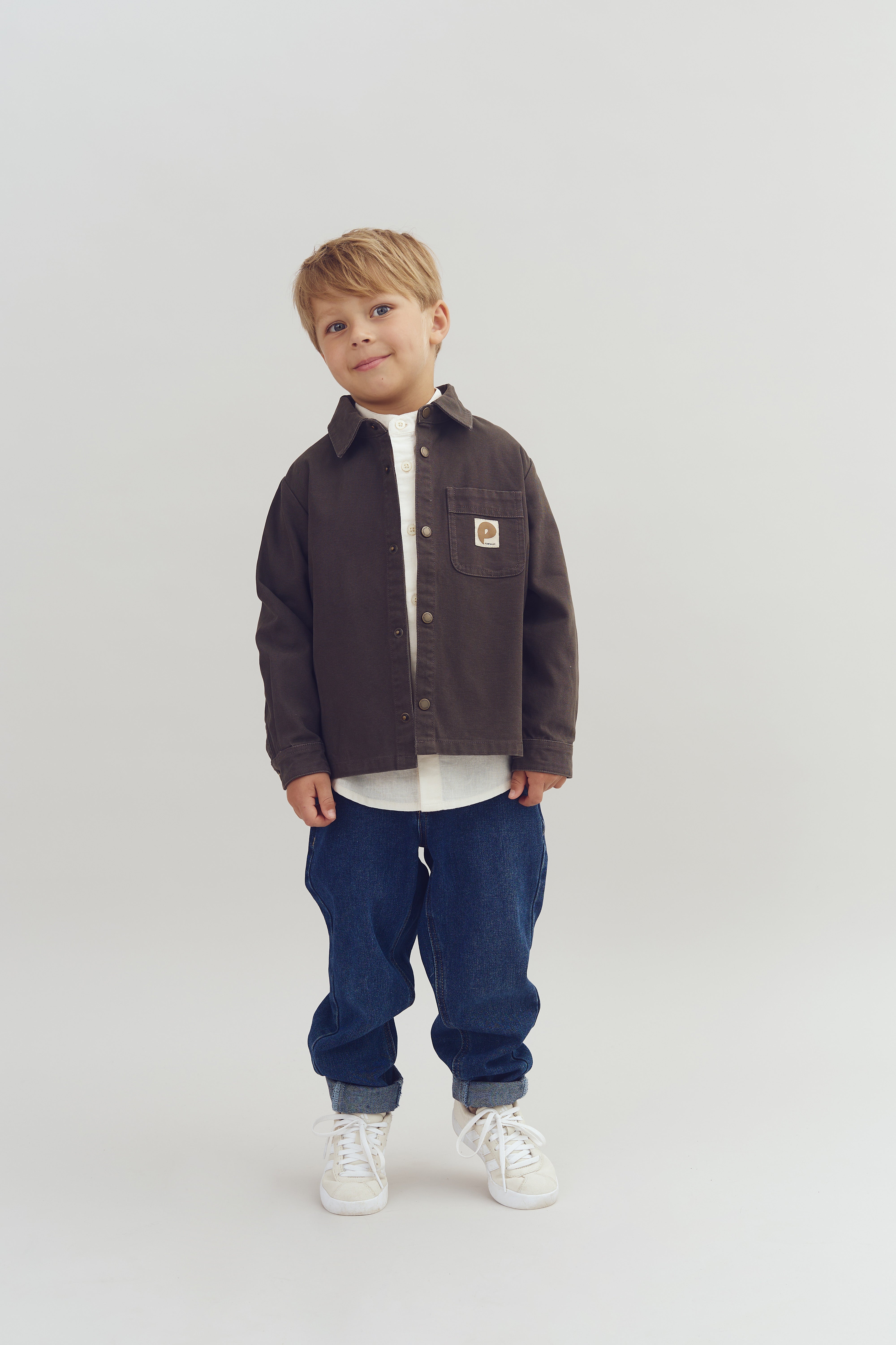Een jongen draagt een donkerbruin jasje met een embleem op de borst, een wit overhemd, blauwe spijkerbroek en witte schoenen, staand tegen een neutrale witte achtergrond.