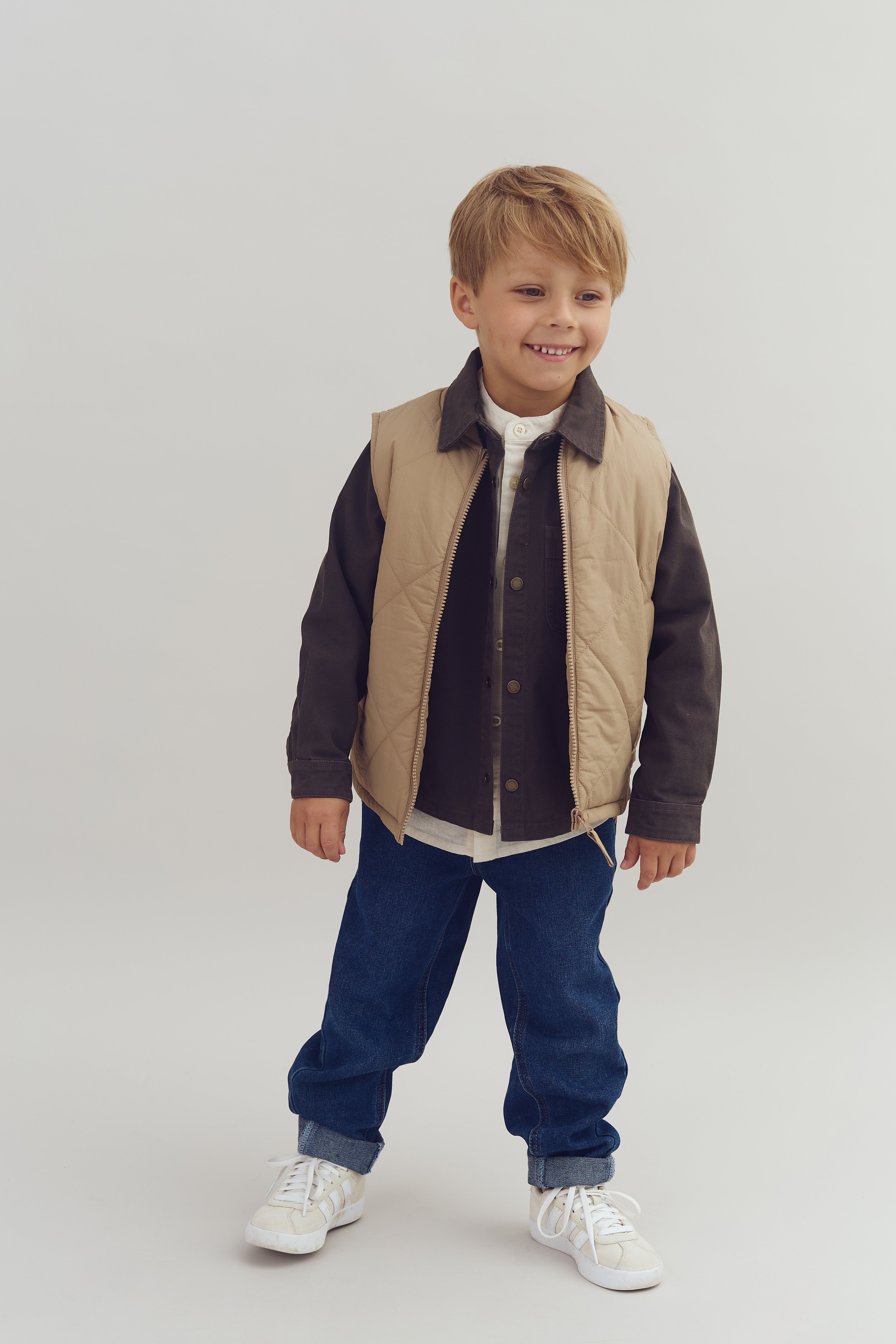 Jongen draagt beige bodywarmer en donker overhemd, staat lachend, met blauwe jeans en witte sneakers in een lichte studio-omgeving zonder tekst.