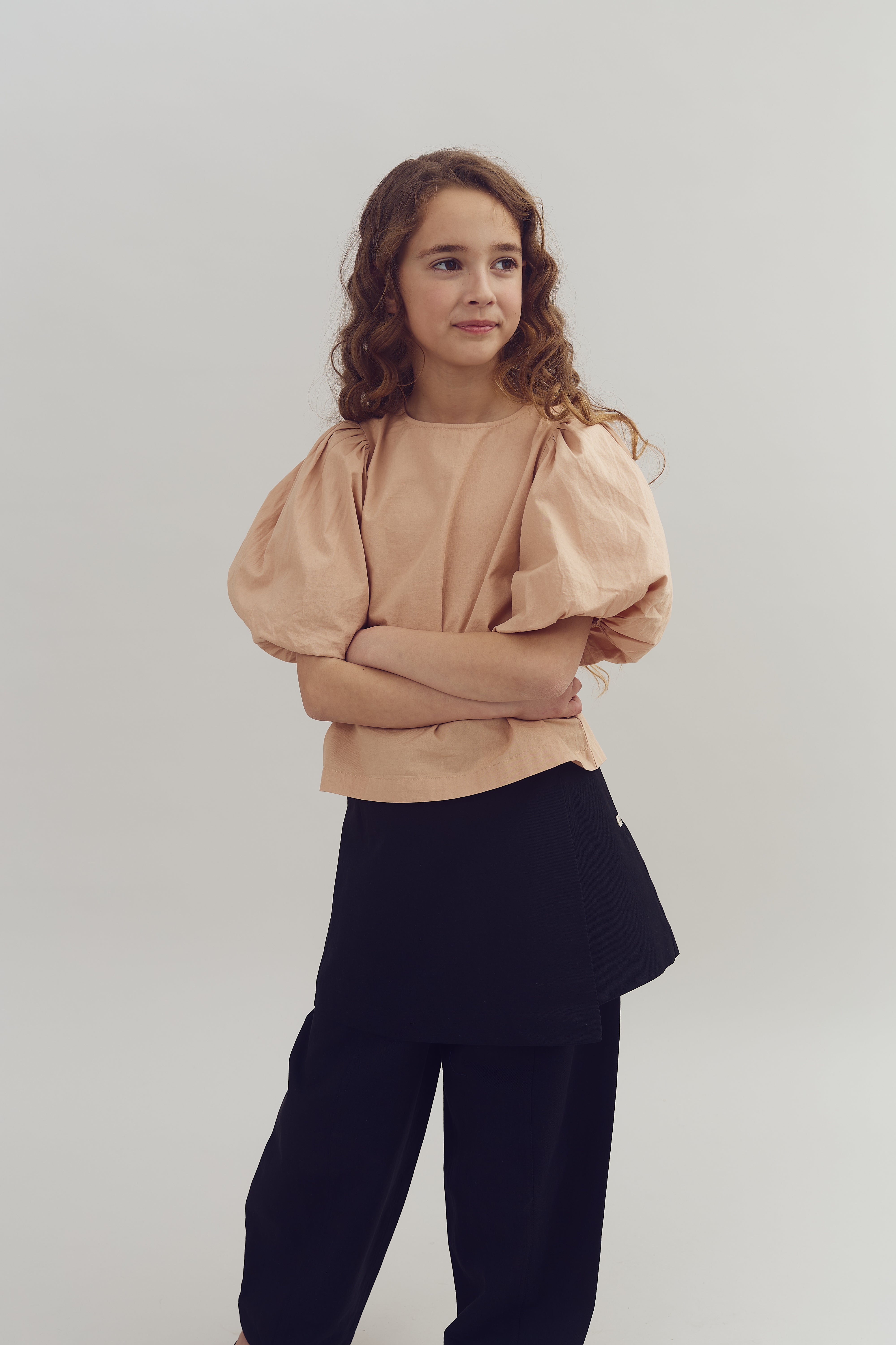 Kind met krullend haar draagt een roze blouse en zwarte broek, staat met gekruiste armen tegen een neutrale, lichtgekleurde achtergrond.